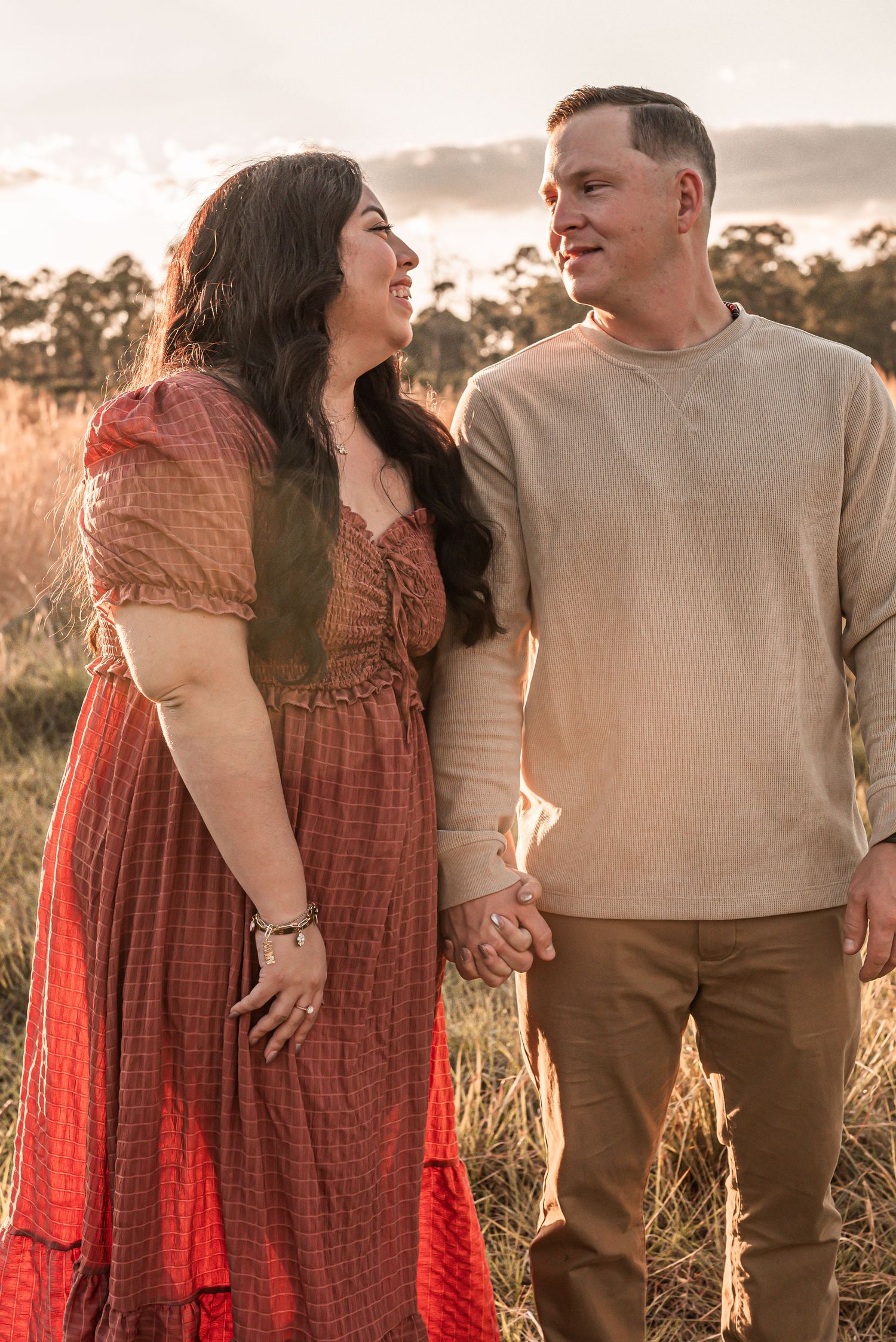 A couple shares a tender moment while holding hands in a field at sunset wearing casual attire.