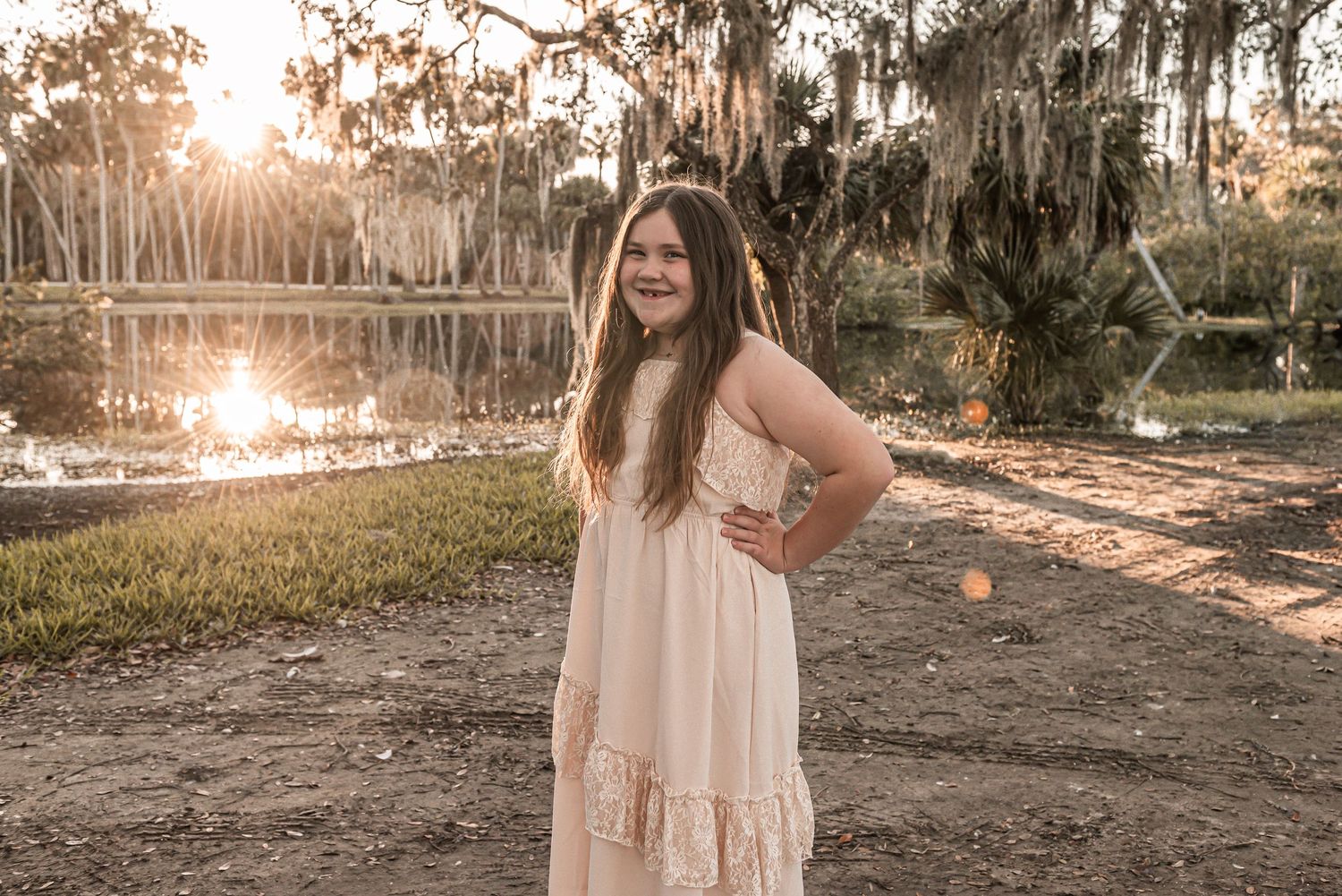 A maxi dress-clad figure stands by a tranquil lake at sunset with Spanish moss hanging from trees in the background.