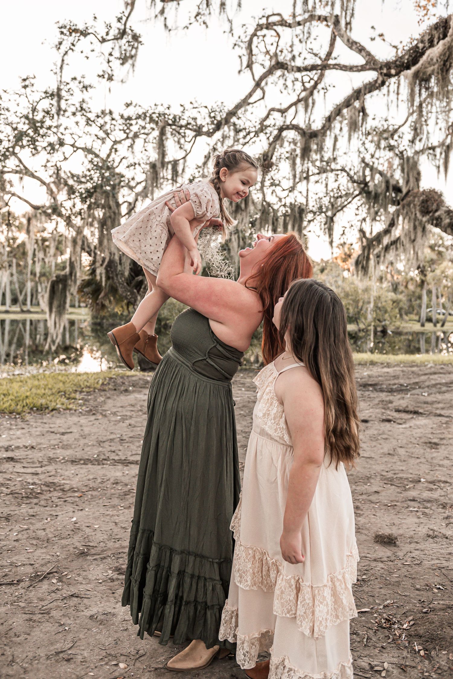 Three people in flowing dresses stand together under Spanish moss draped trees in a dreamy outdoor setting at sunset.
