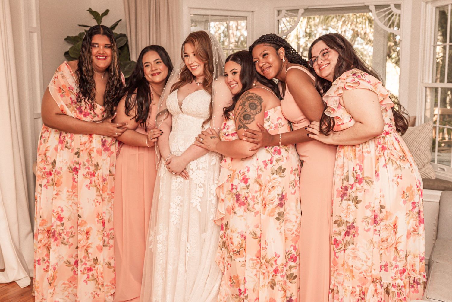 Group wearing pink floral and solid bridesmaid dresses pose with bride in white gown at wedding celebration.
