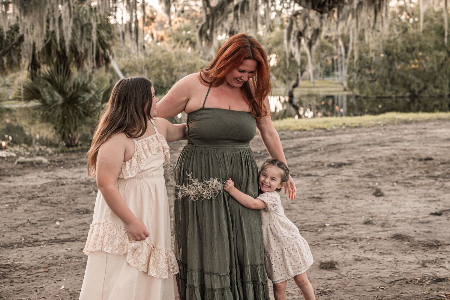 Two children in cream-colored dresses stand with a figure wearing an olive green maxi dress in a rustic outdoor setting.