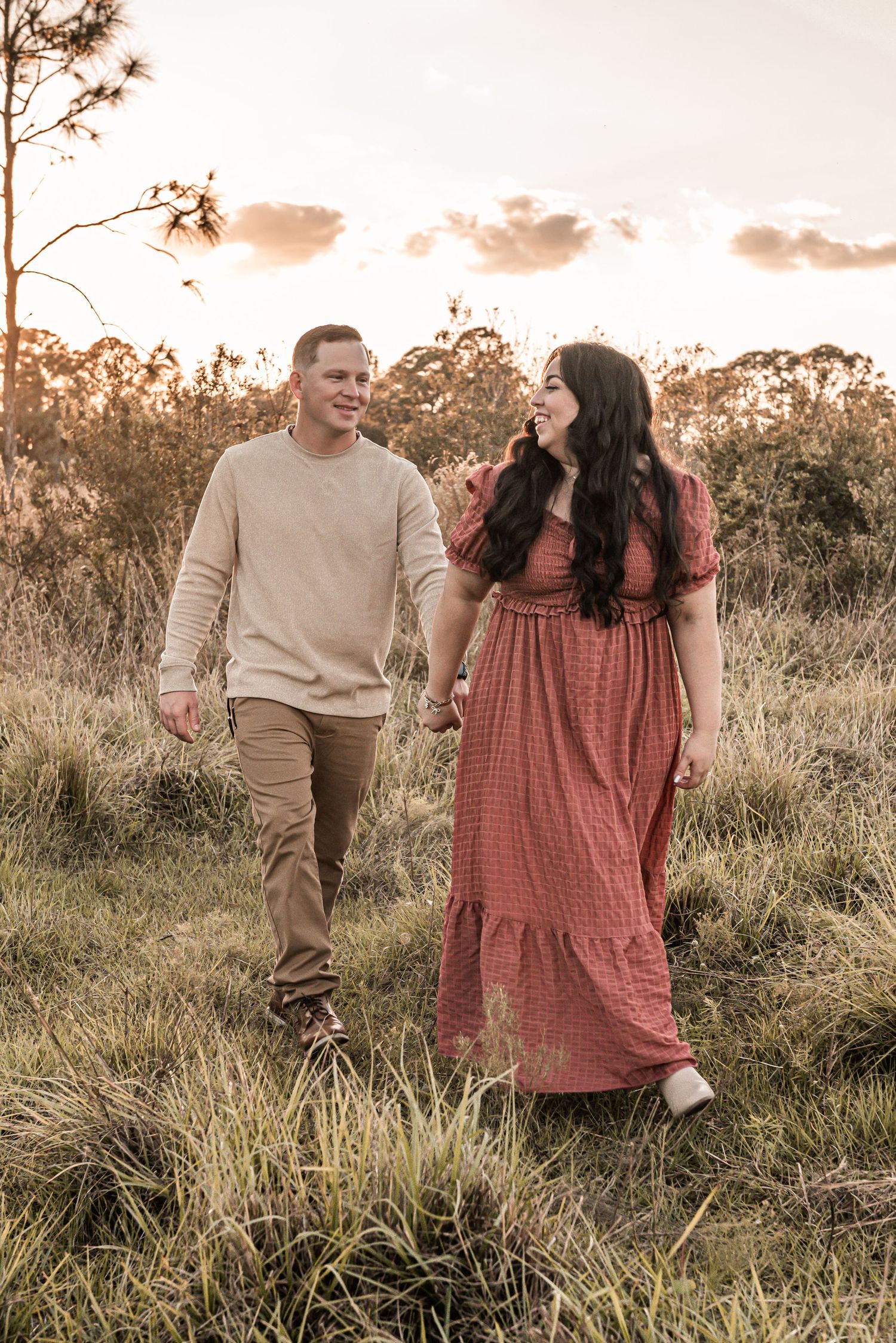 A couple walks together through tall grass in a golden field at sunset, sharing a joyful moment.