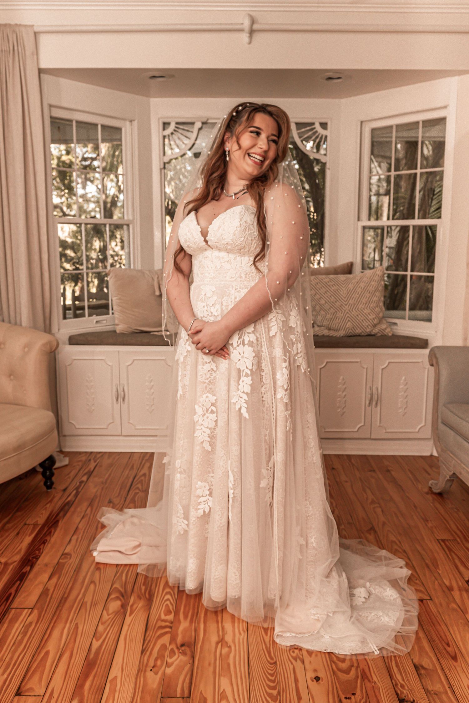 Elegant bride wearing a flowing lace wedding gown poses in front of a bay window in a classic interior setting.