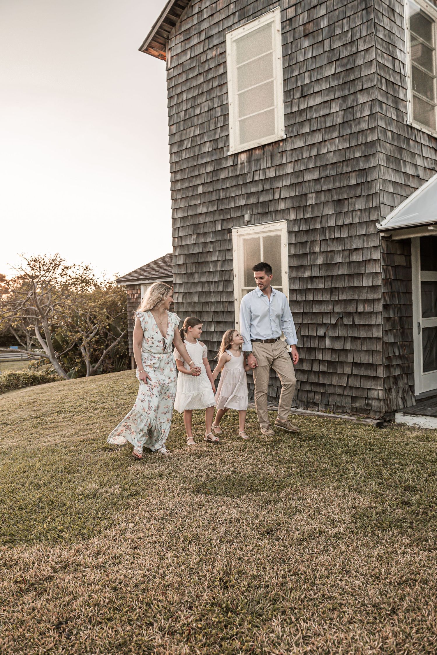 Rustic shingle-sided house with figures walking in yard at sunset with soft golden light.