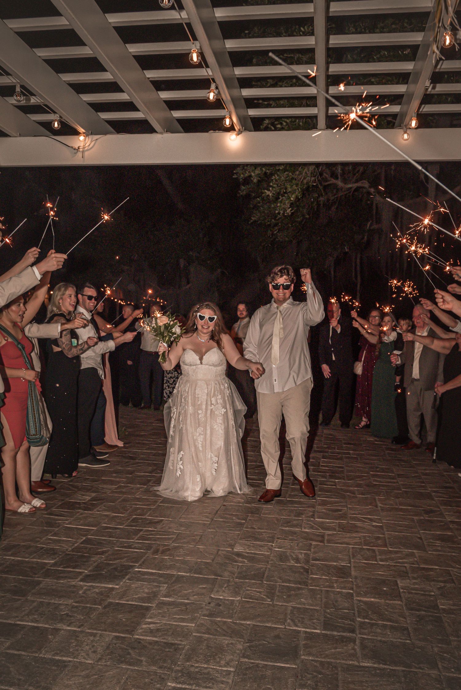 Wedding couple celebrates their sparkler exit during nighttime reception on an outdoor stone patio under string lights.