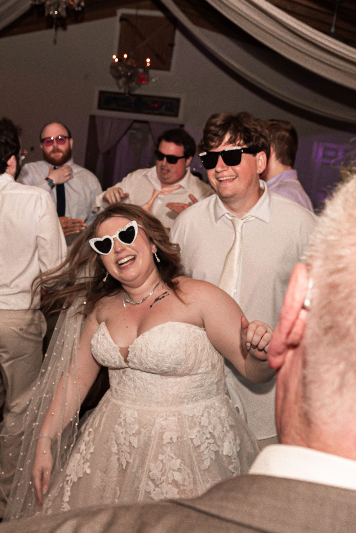Cheerful bride in lace dress and sunglasses dances with wedding guests wearing white shirts at indoor reception party.