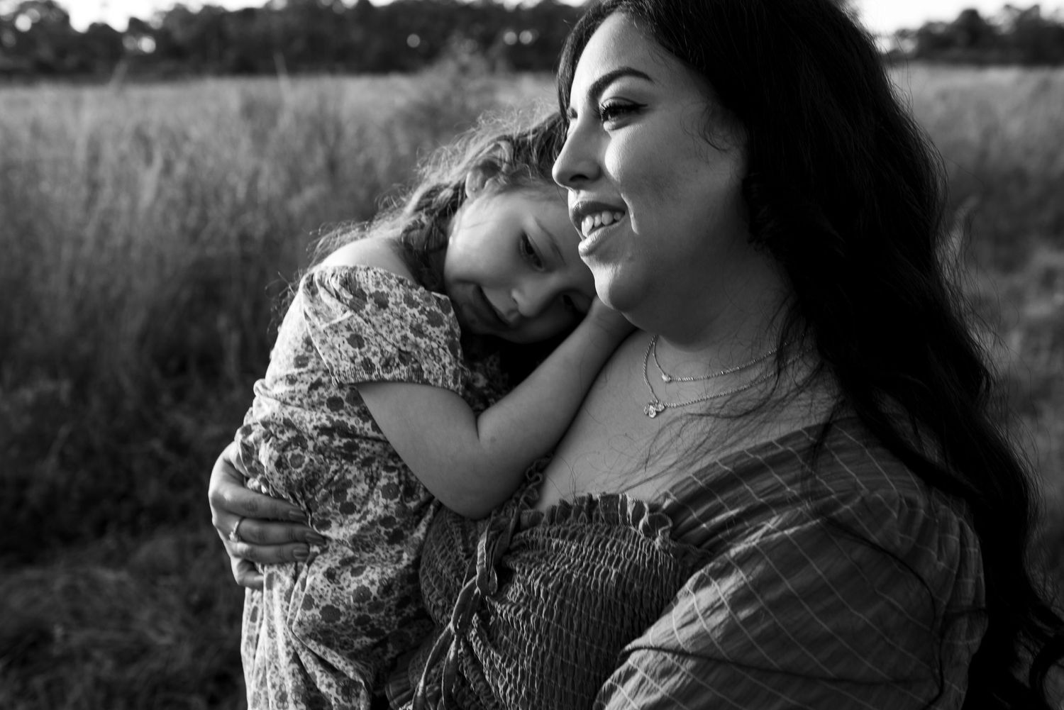 Black and white portrait showing tender moment between parent and child outdoors in a grassy field at sunset.