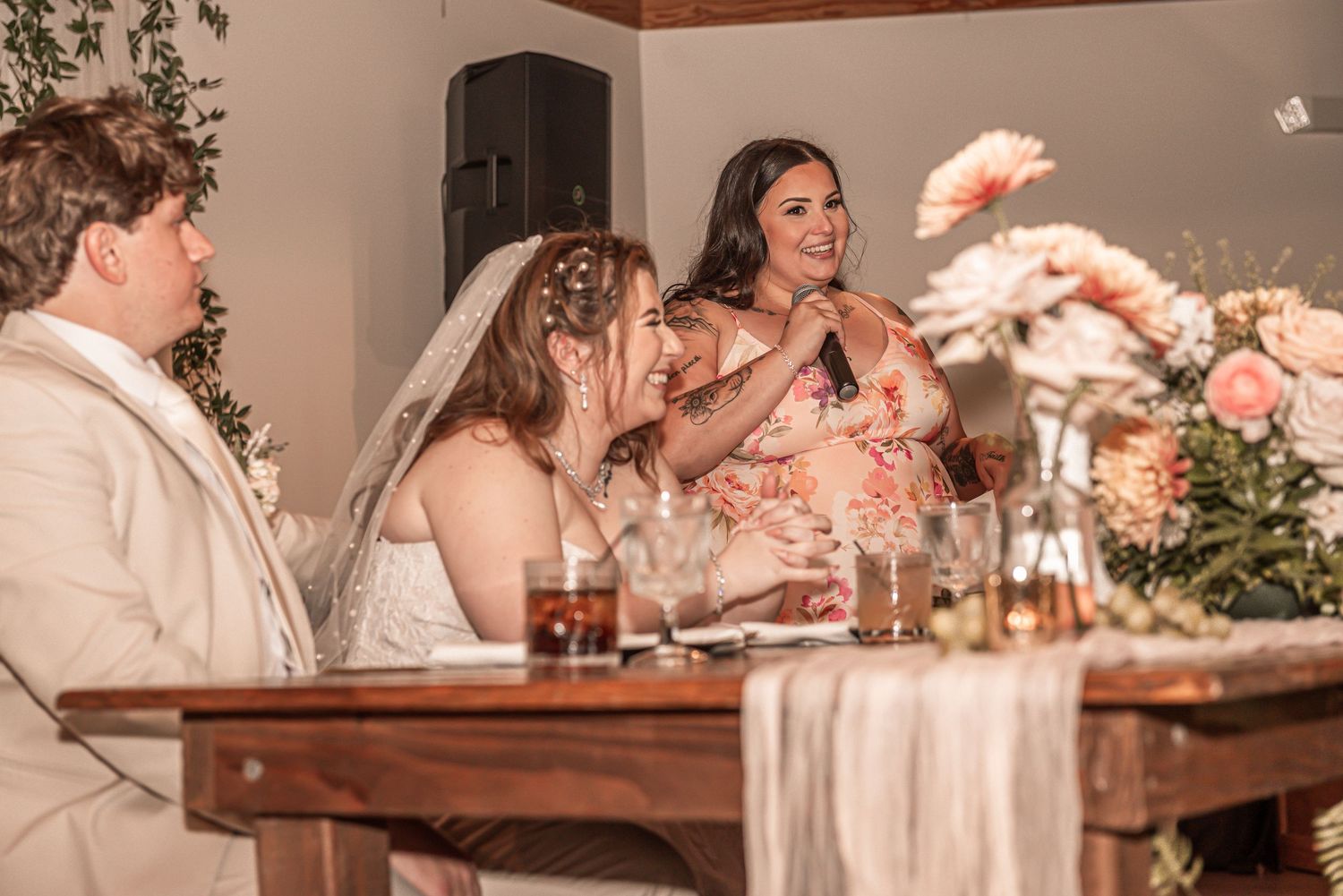 People gather at a wedding reception dinner table decorated with floral centerpieces and candles in warm lighting.