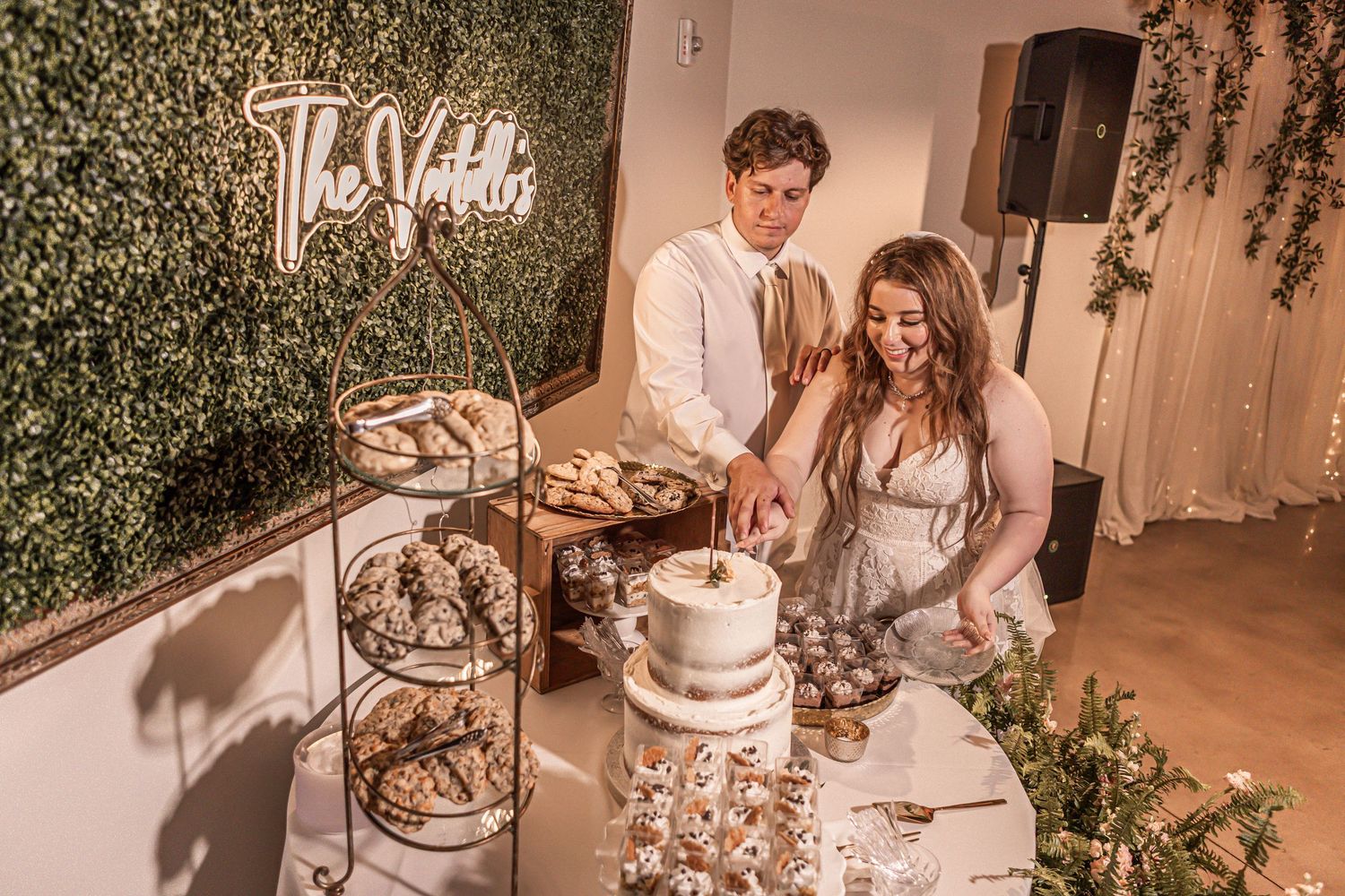A couple cuts their elegant white wedding cake together at a reception with a green floral wall backdrop and dessert table.