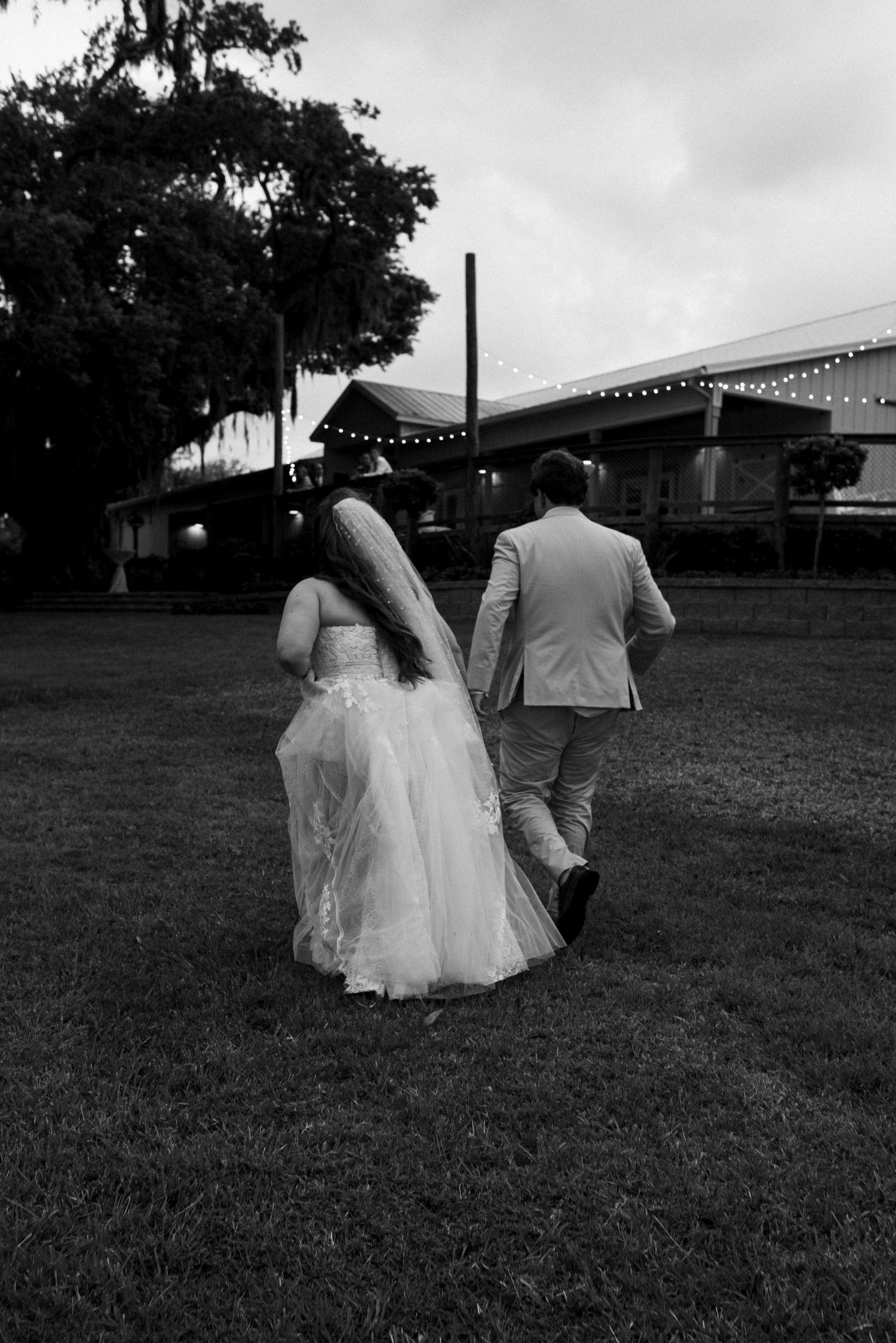 A black and white wedding photograph of a couple walking on grass with string lights and trees visible in the background.
