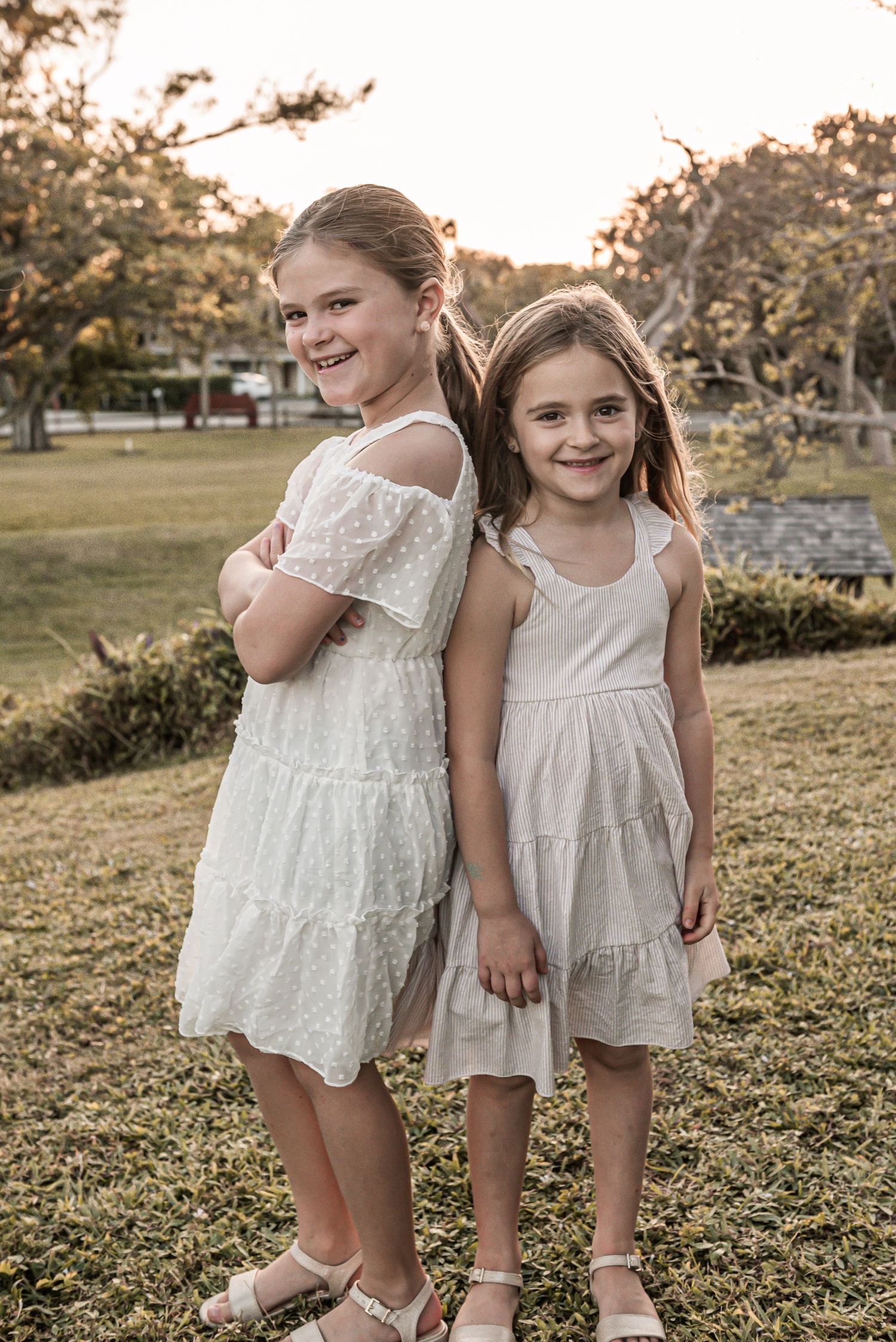 Two young girls wearing white summer dresses pose together at sunset in a grassy park setting.