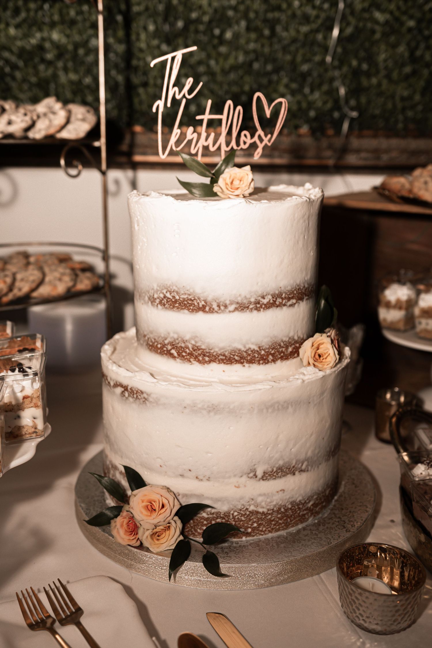 A rustic three-tier semi-naked wedding cake decorated with peach roses and a gold script topper on a dessert table.