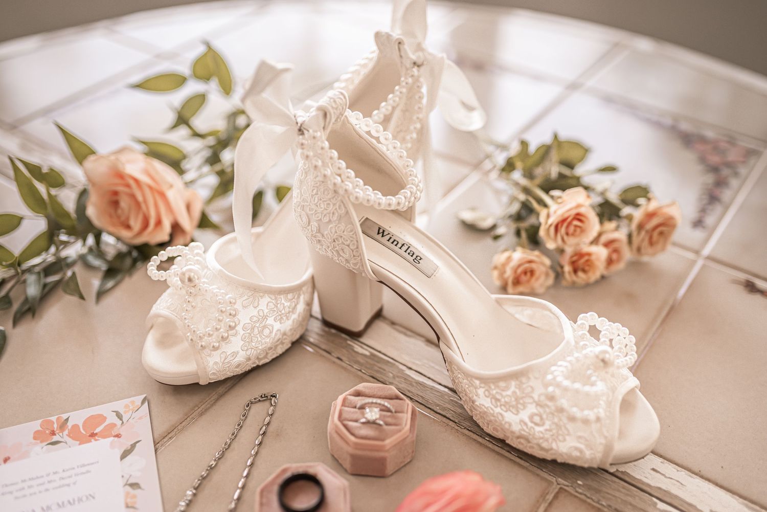 Elegant white lace bridal shoes with block heels displayed alongside delicate peach roses and wedding accessories.