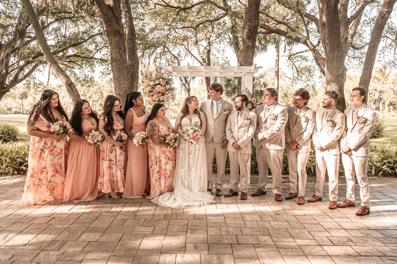 A wedding party poses outdoors wearing peach and beige tones on a brick patio under shade trees.