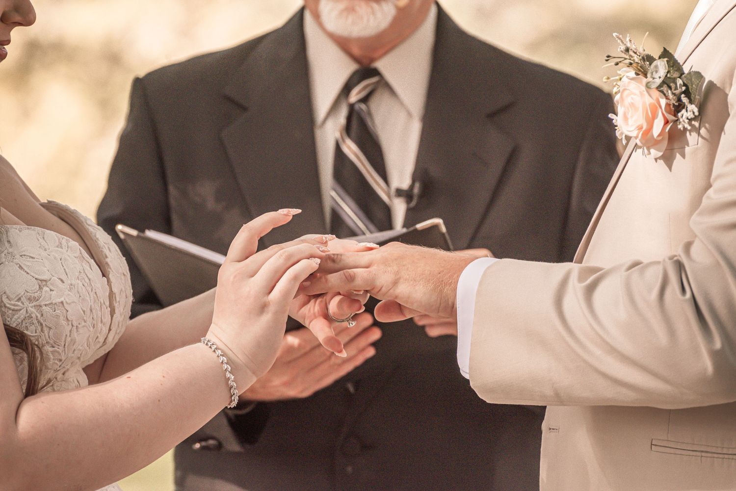 Close-up of hands exchanging wedding rings during ceremony with officiant in background.
