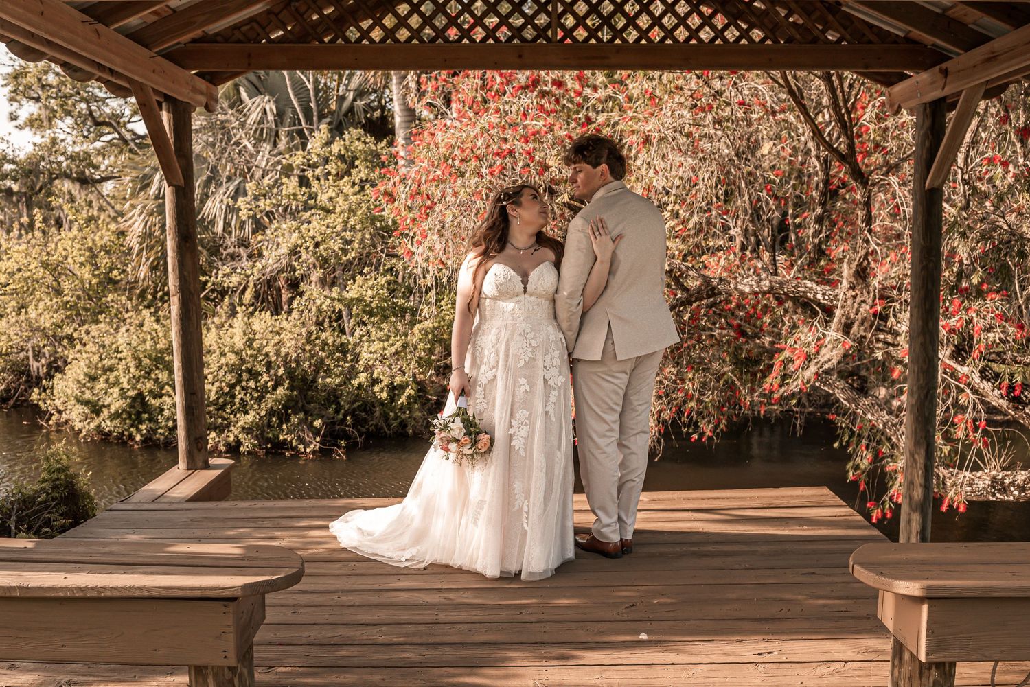 A couple stands together under a wooden gazebo on their autumn wedding day surrounded by fall foliage.