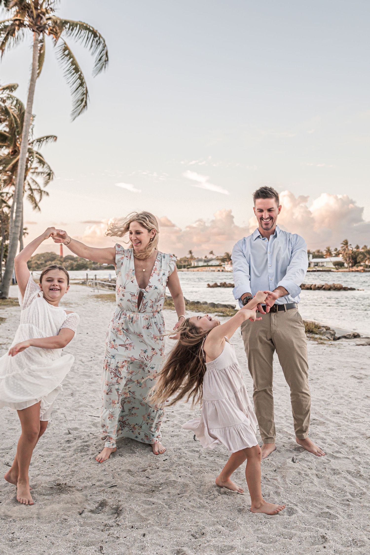 A family plays and dances together on a tropical beach at sunset with palm trees and ocean in the background.
