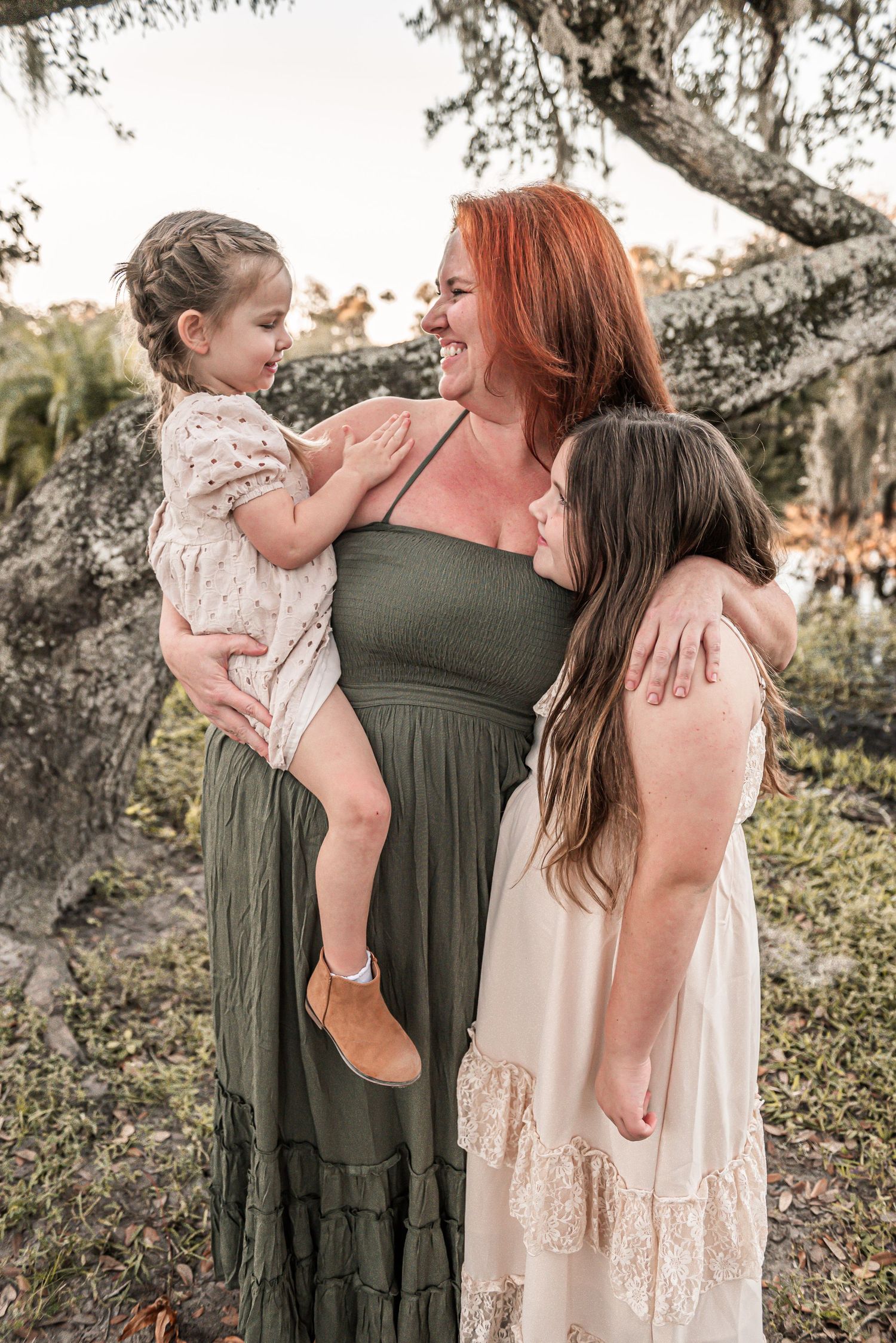 A tender family moment outdoors shows a woman in an olive dress embracing her children in cream-colored dresses under a tree.