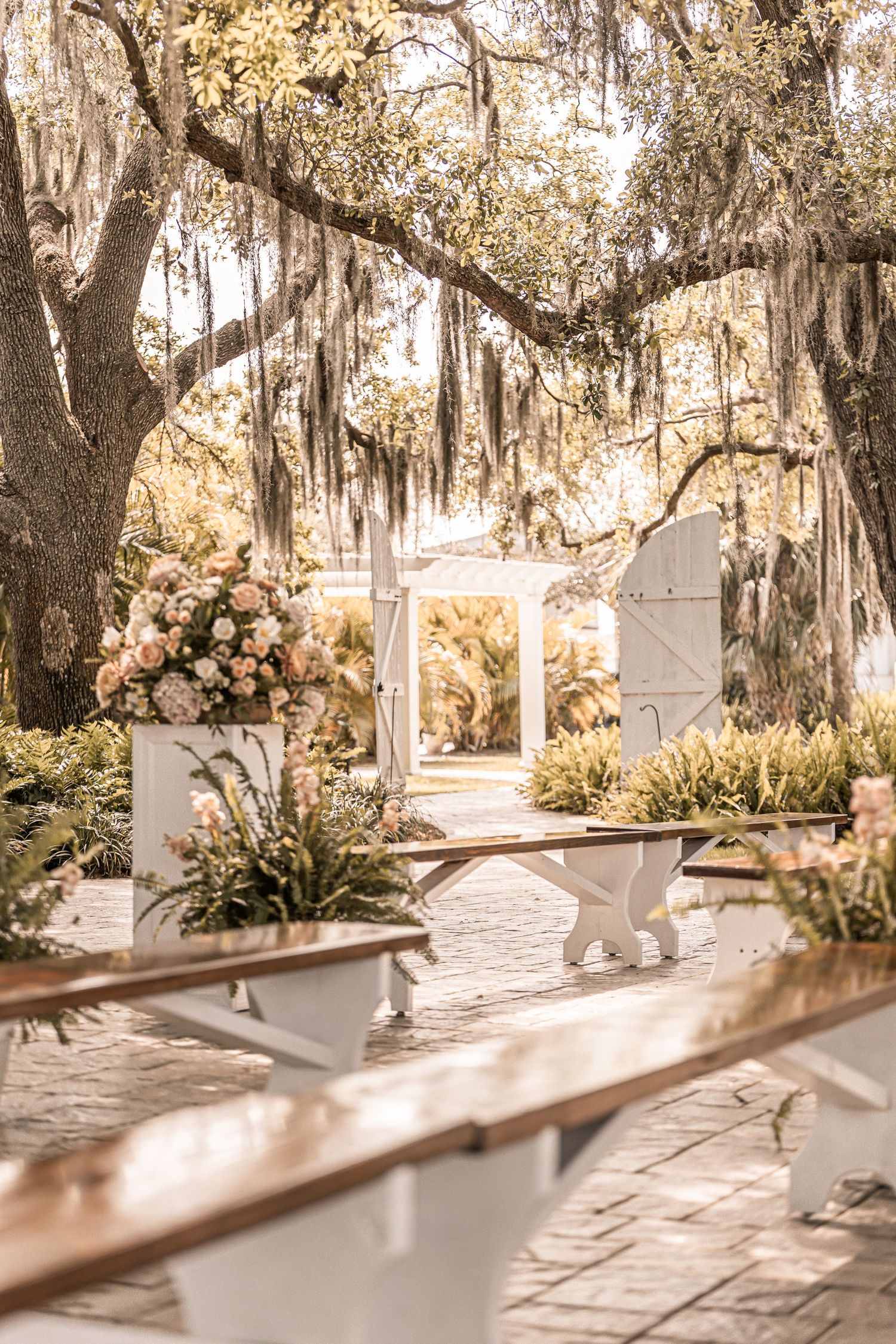 A romantic garden pathway with hanging Spanish moss and white floral arrangements leads to an elegant white archway.