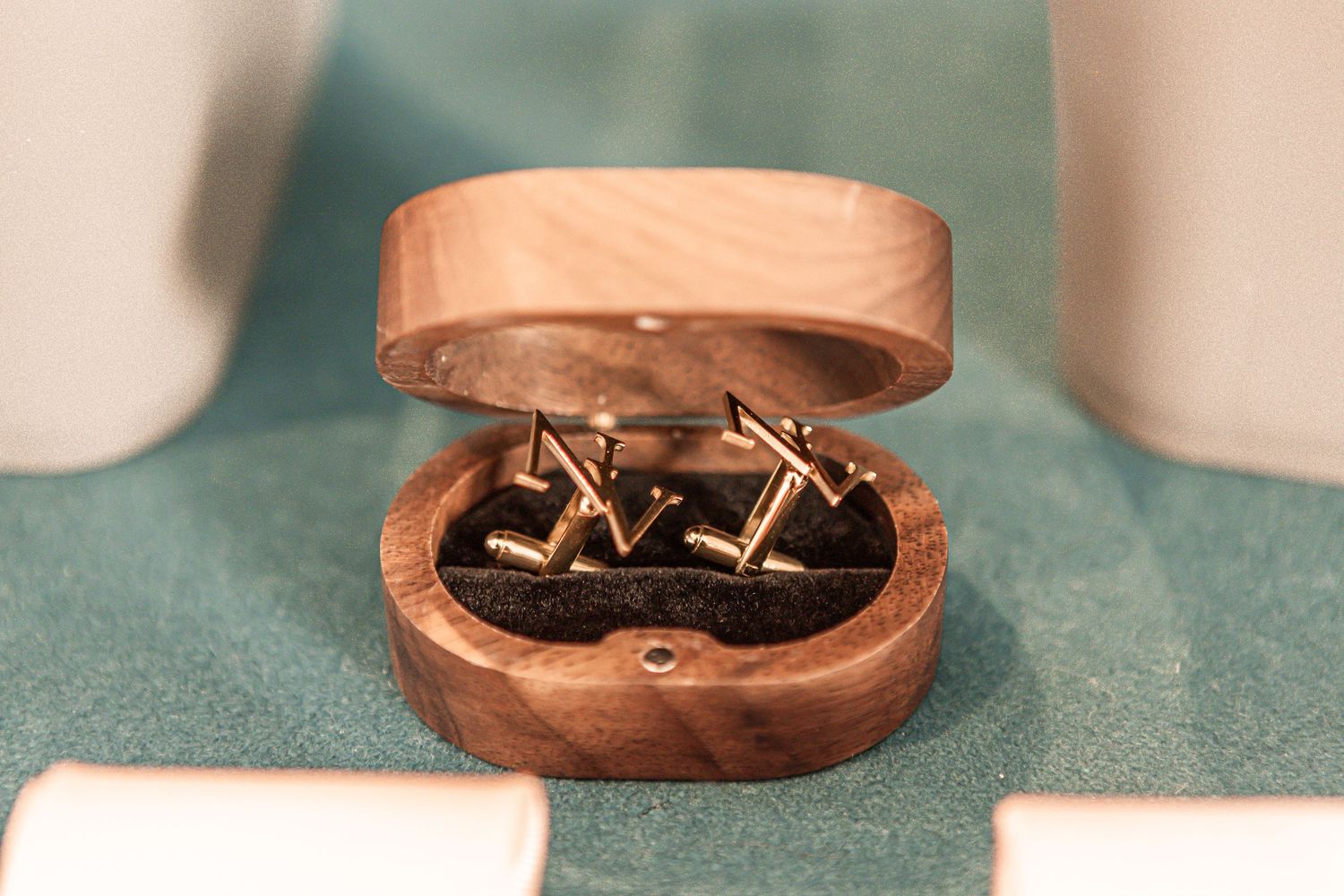A pair of gold cufflinks displayed in a wooden ring box on a teal green surface.