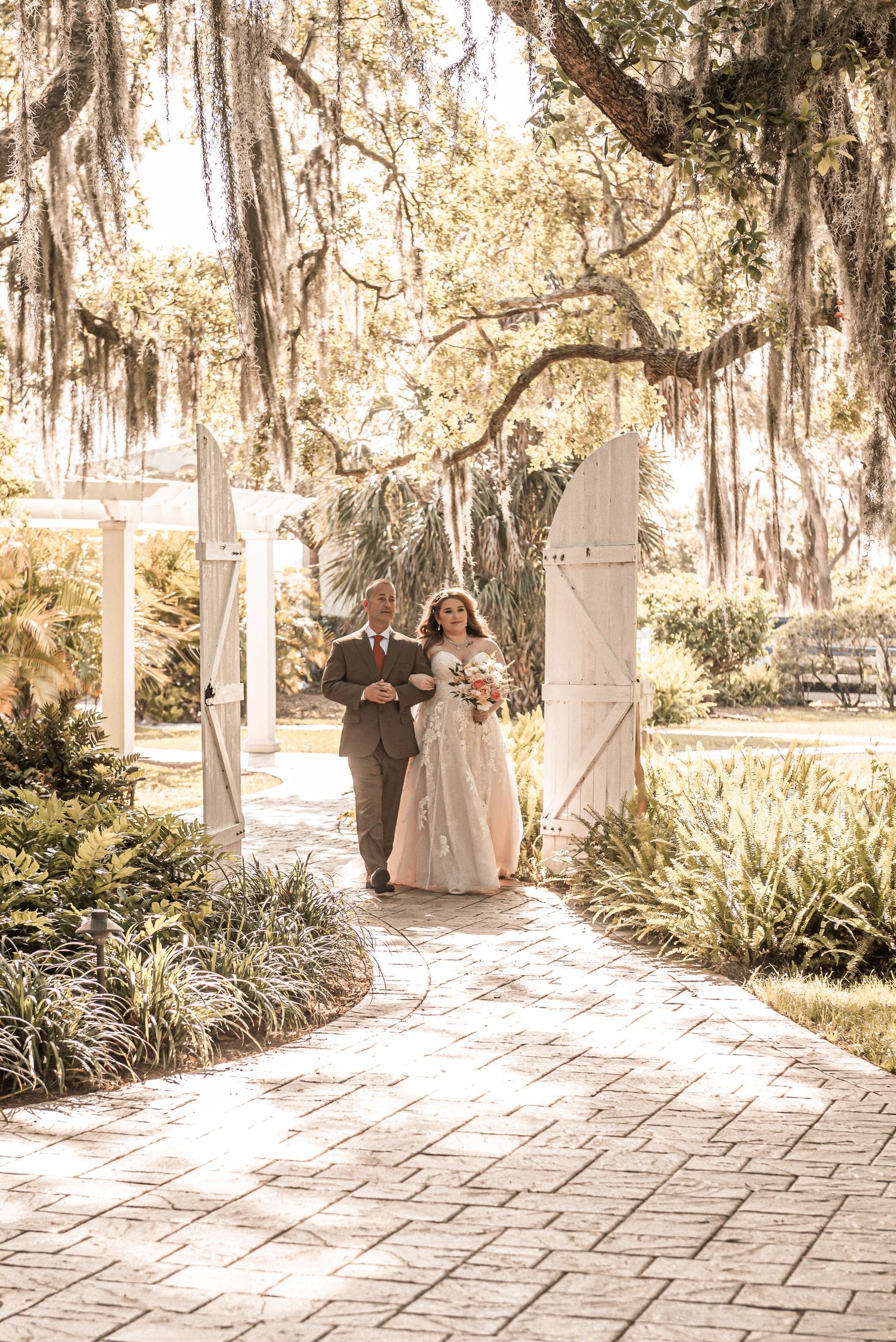 A romantic wedding ceremony path under Spanish moss draped oak trees and white columns on a sunny garden day.