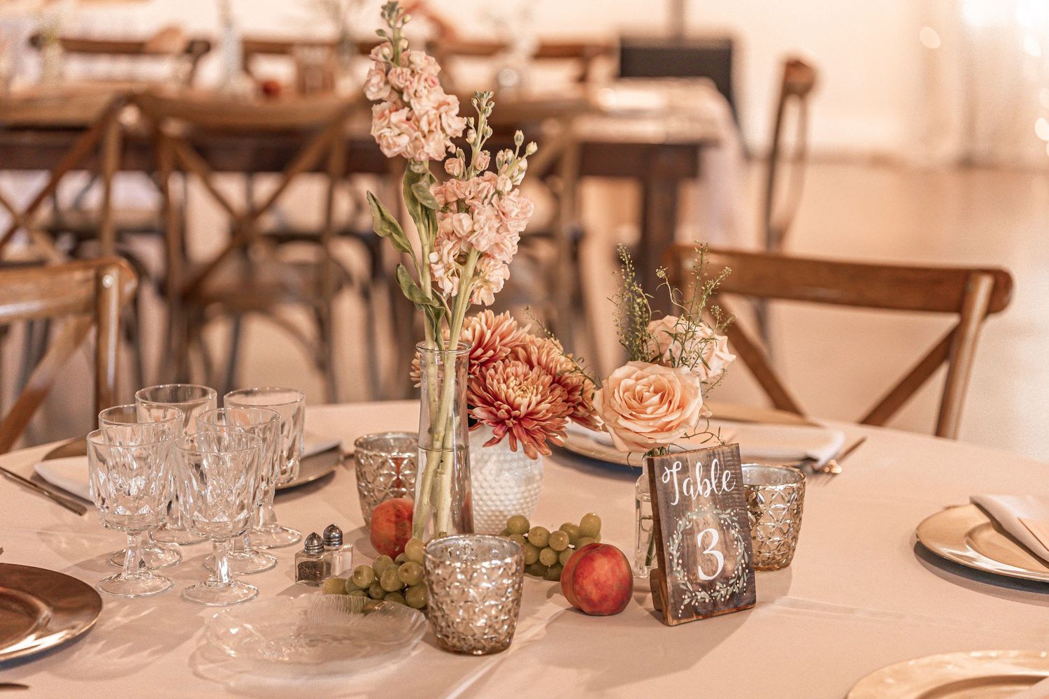 Rustic wedding table setting with delicate floral centerpiece, mercury glass votives, and vintage glassware on white tablecloth.