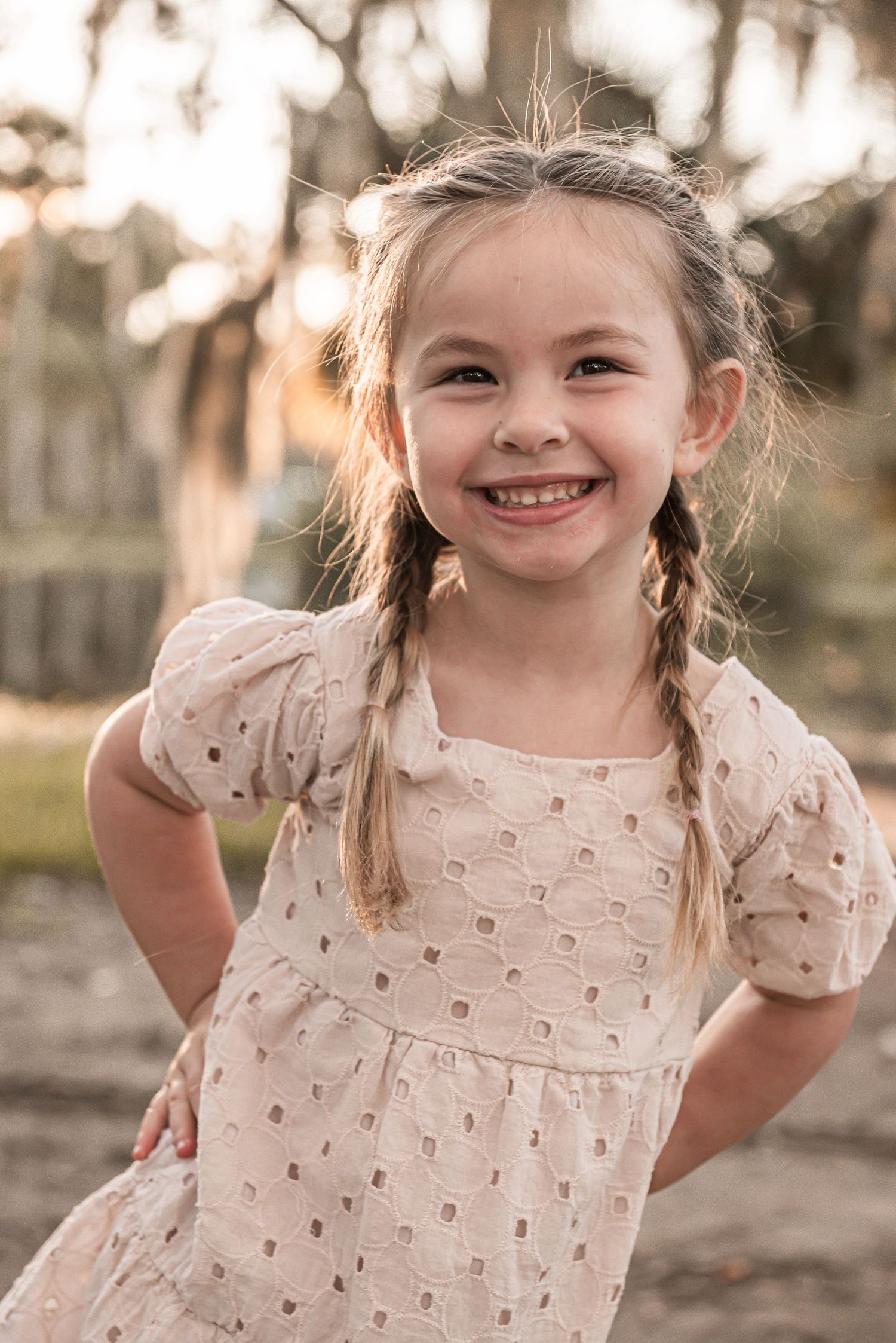 A joyful candid outdoor portrait showing a child wearing a cream-colored dress with braided hair in natural lighting.