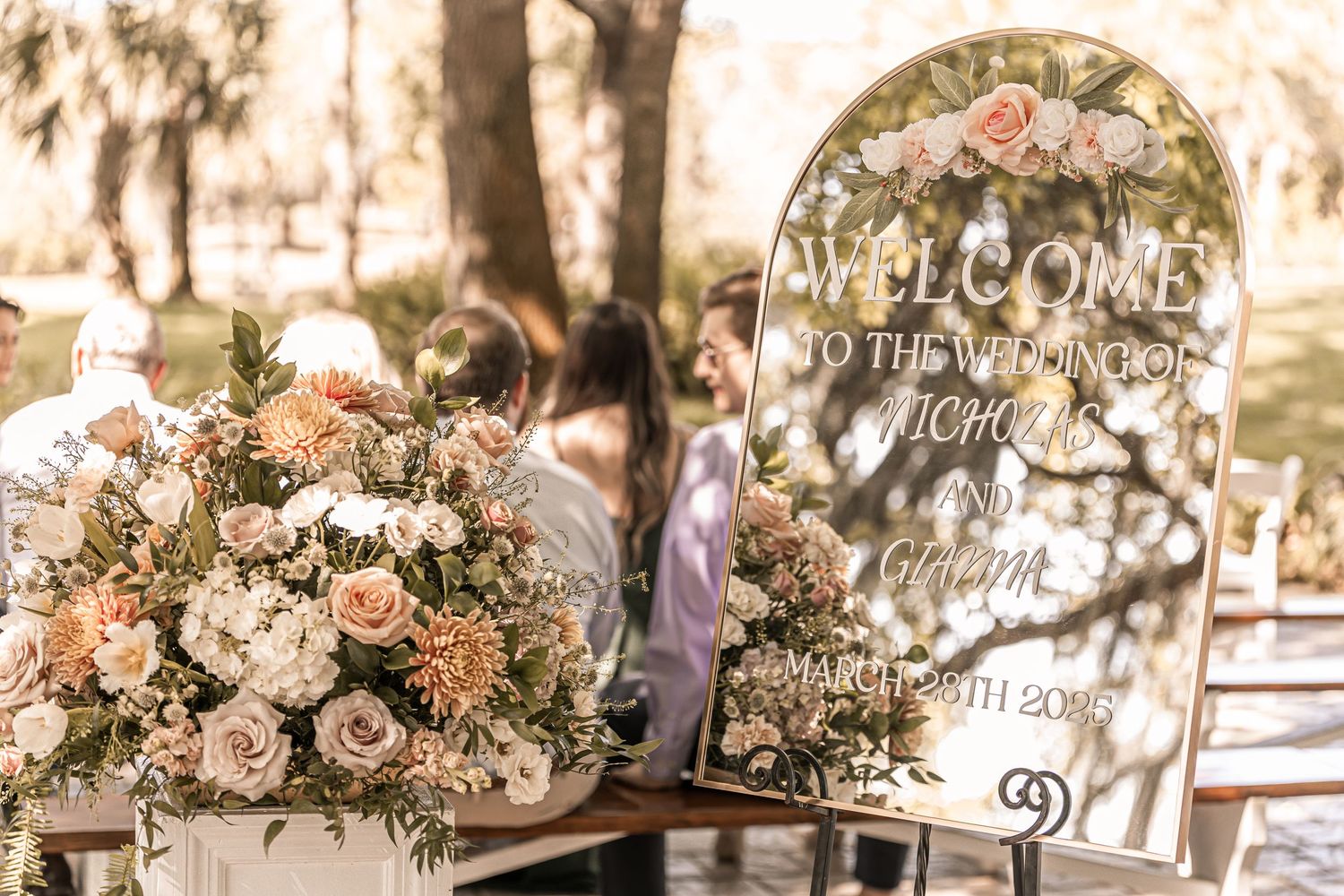 Welcome sign and floral arrangement with pink and white roses and greenery at an elegant outdoor wedding venue.