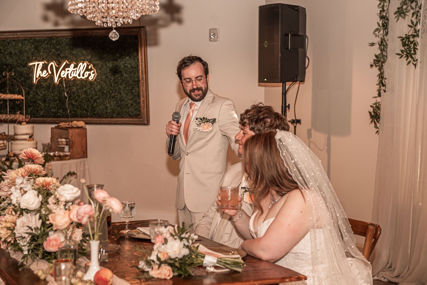 A wedding reception scene showing guests at a dinner table with beautiful floral arrangements and decorative lighting.