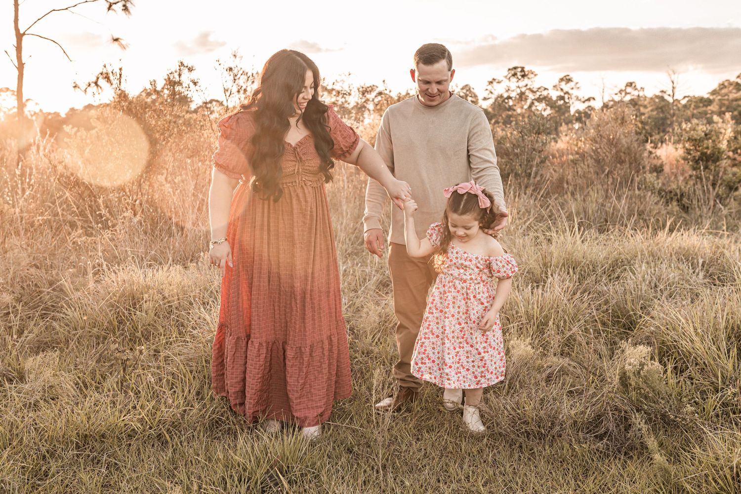 A family walks together in a golden sunlit meadow at sunset wearing coordinated pink and neutral outfits.