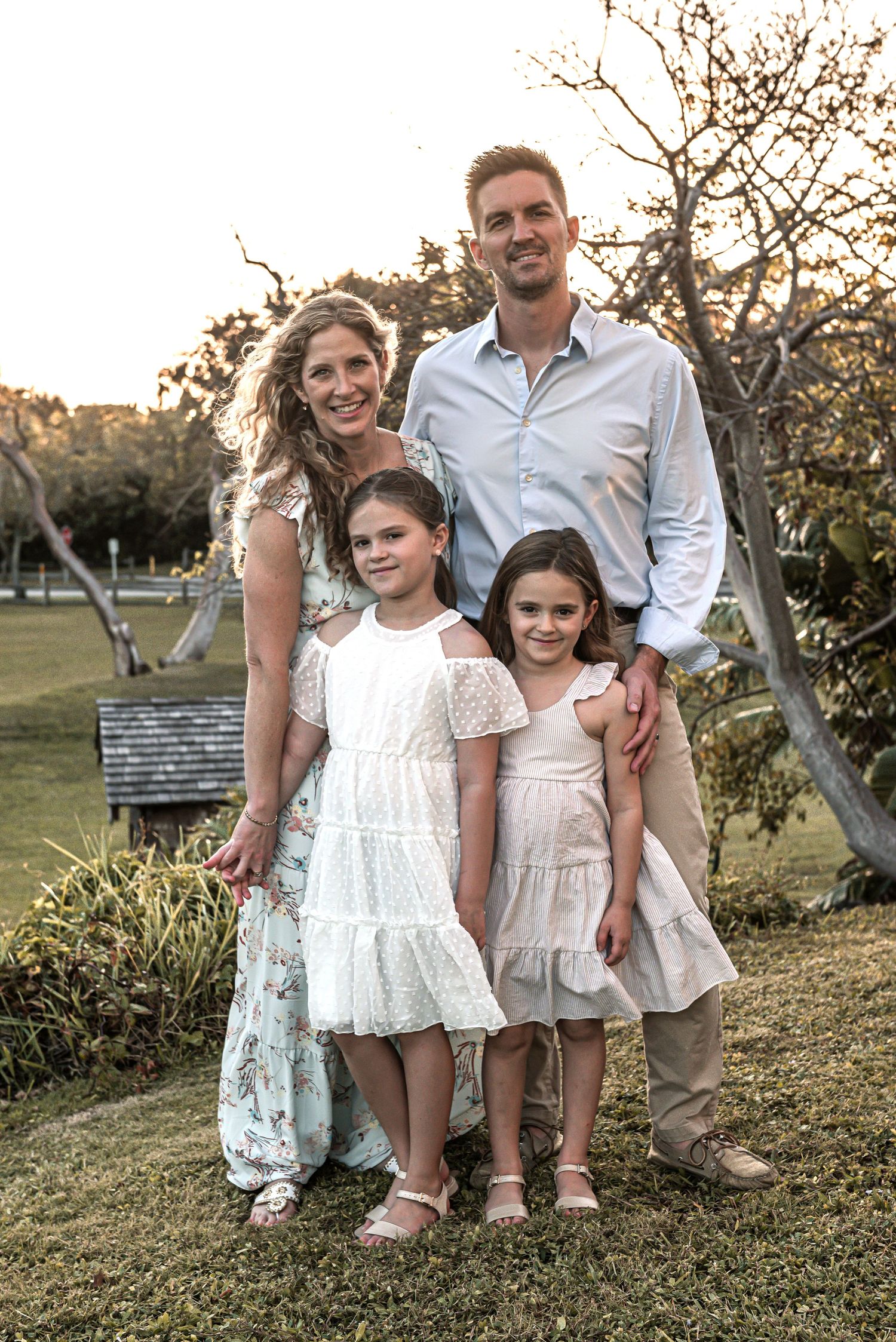 A family poses outdoors at sunset in matching white outfits on a grassy hill with trees in the background.
