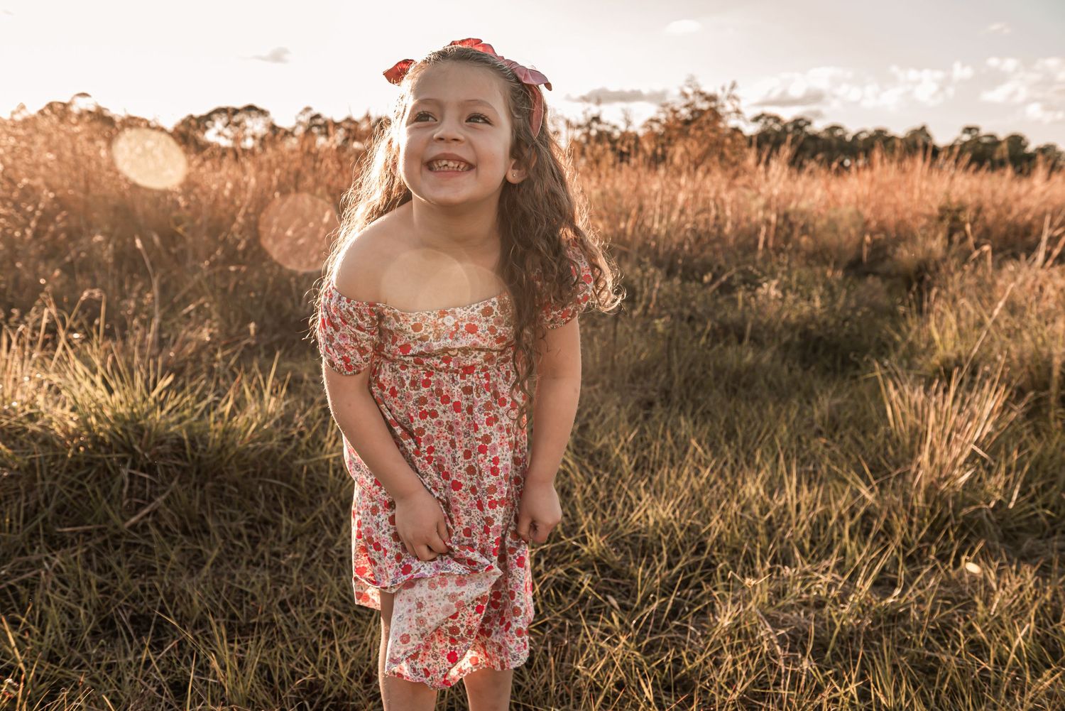 Young child wearing a floral dress runs through a sunlit field of tall grass at golden hour.