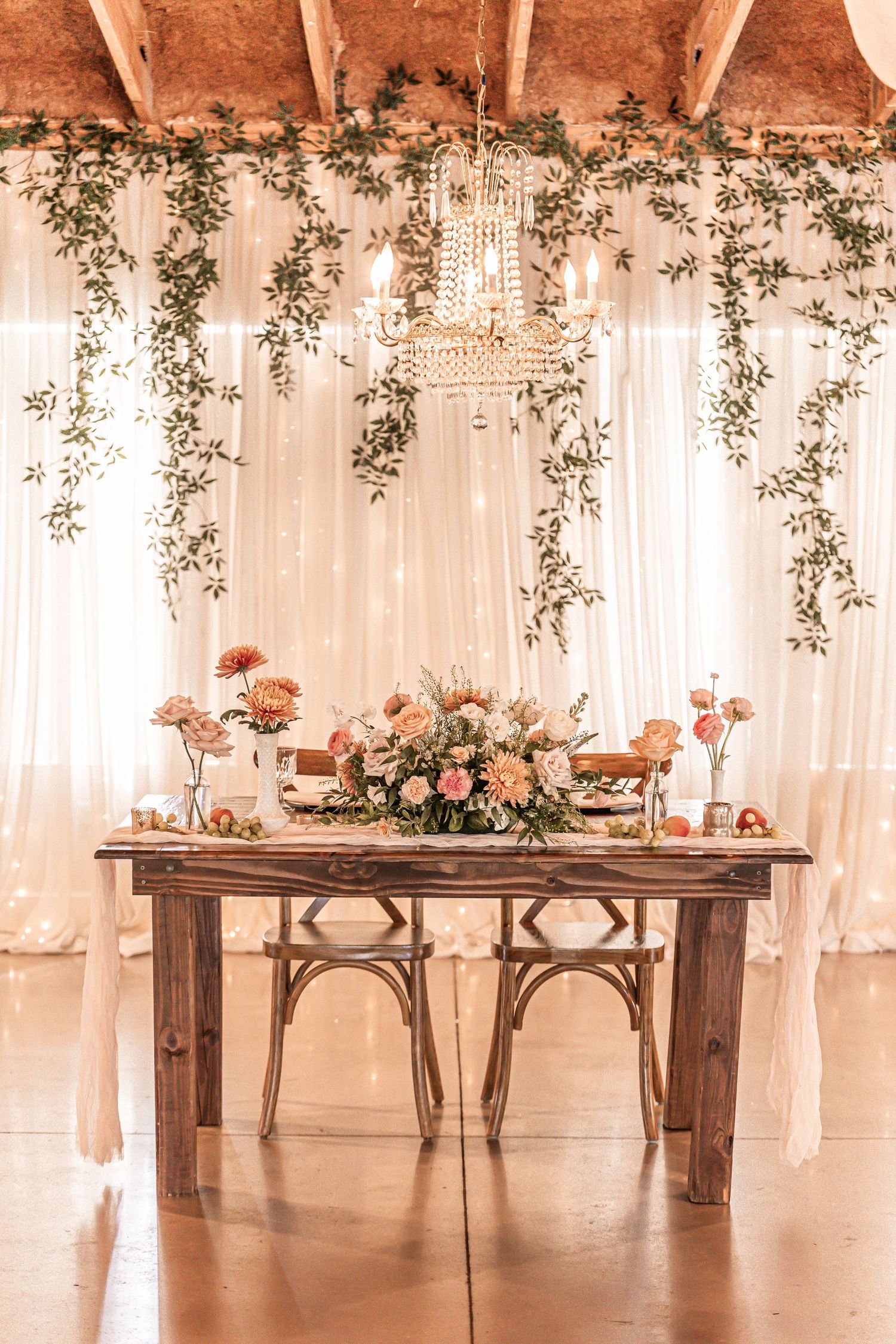 Elegant wedding sweetheart table setup with crystal chandelier and cascading greenery against white drape backdrop.
