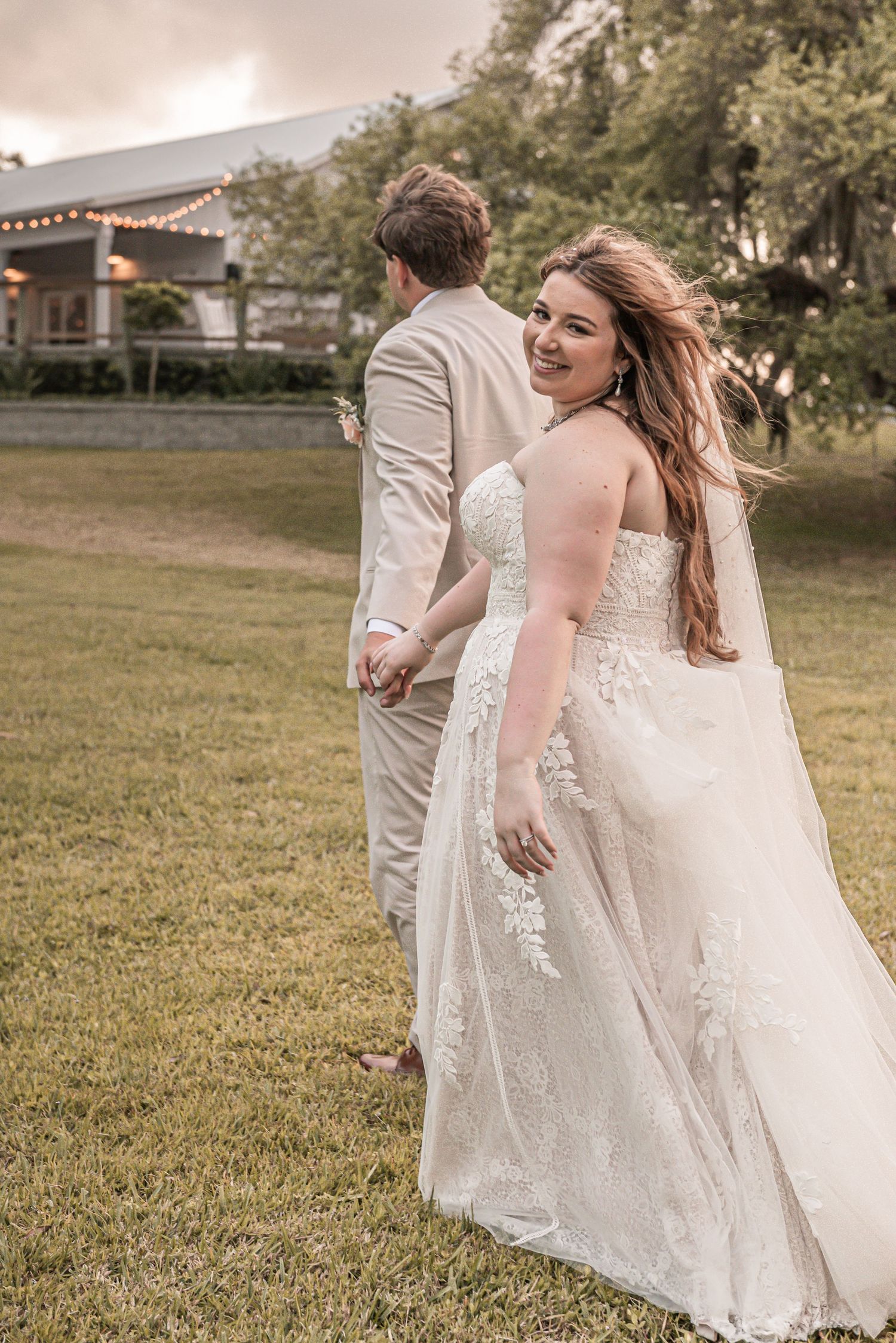 A wedding couple stands in a grassy field with string lights and buildings visible in the background.