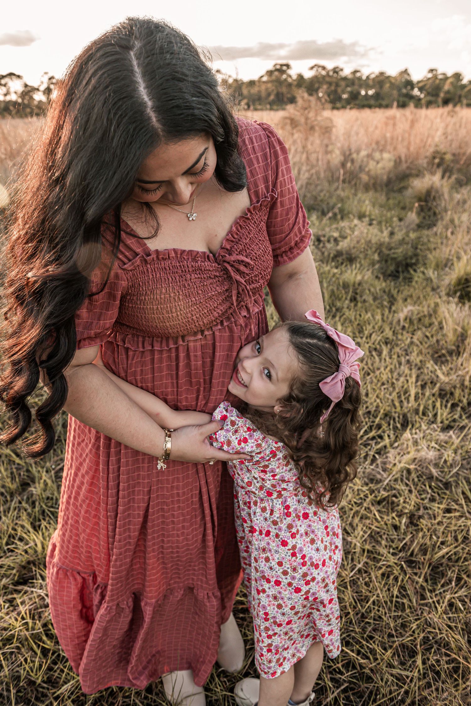 A tender outdoor moment captured between a woman in a rust-colored dress and young child in floral print dress at sunset.