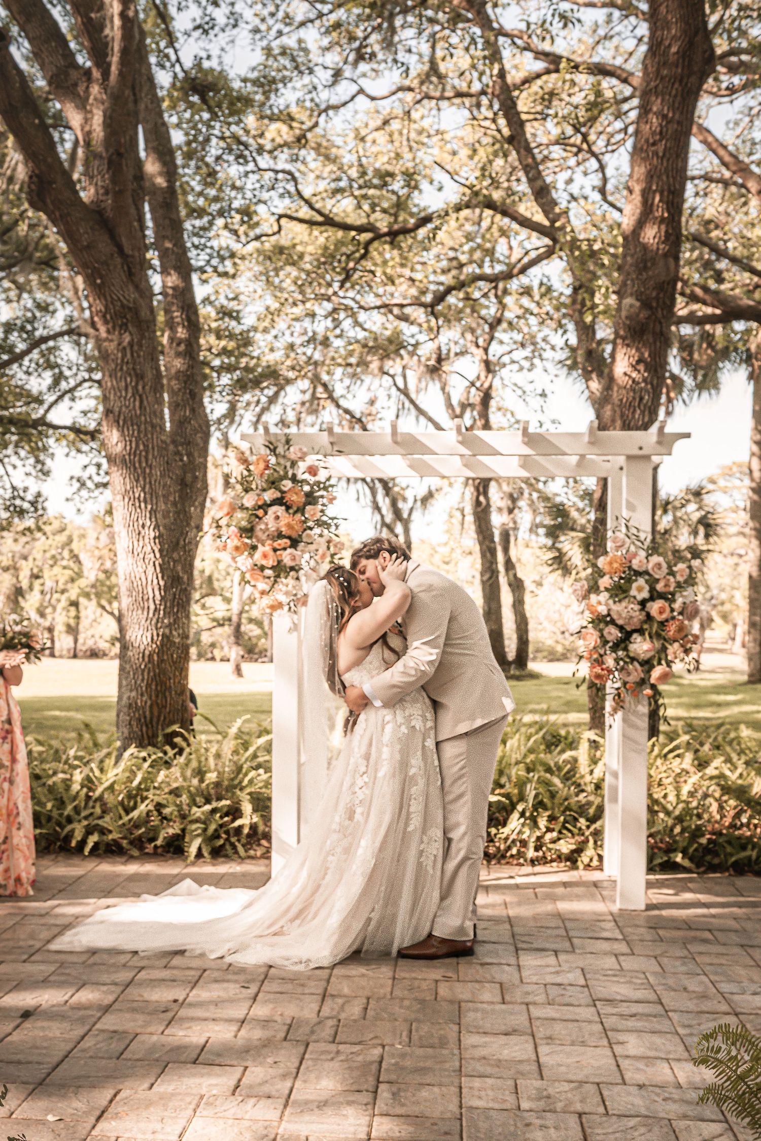 A couple shares a tender moment under a floral-adorned white wedding arbor on a stone patio surrounded by oak trees.