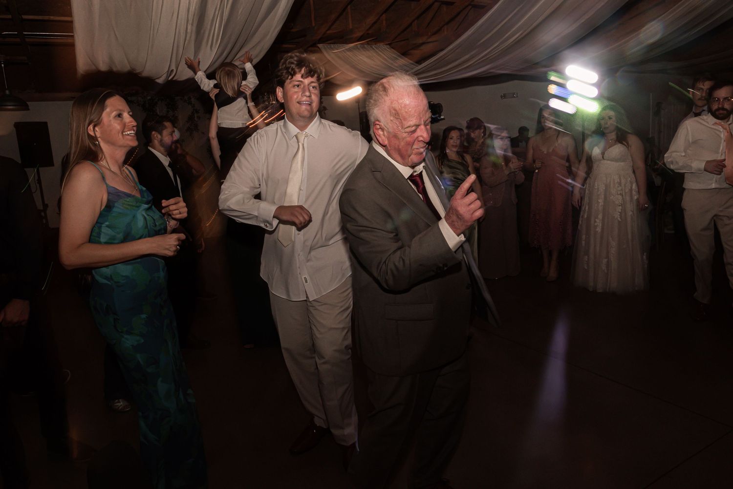 Wedding guests enjoy dancing and socializing on a dimly lit dance floor at an indoor reception.