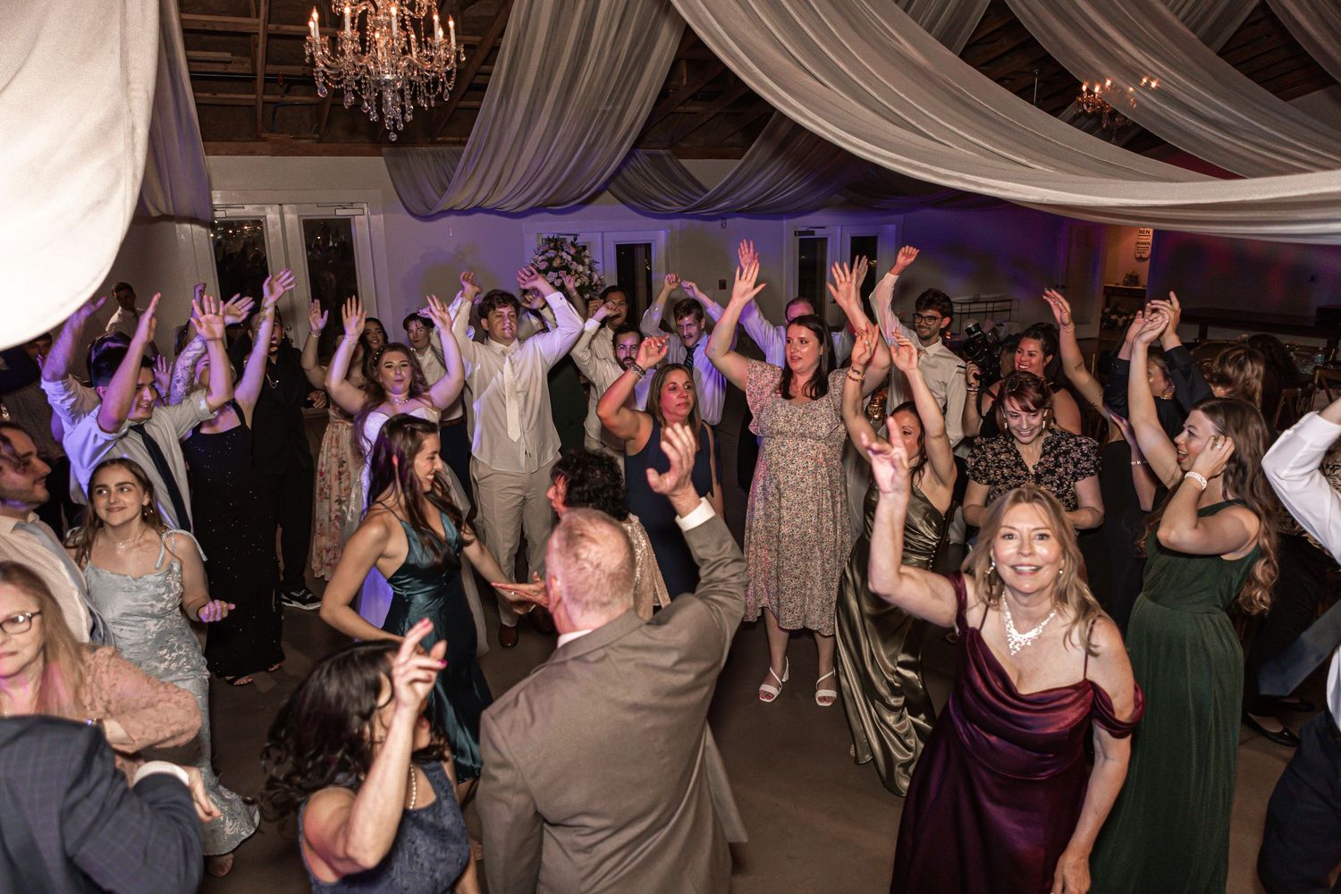 Wedding reception party guests dancing and celebrating on a dance floor under draped fabric and chandeliers.