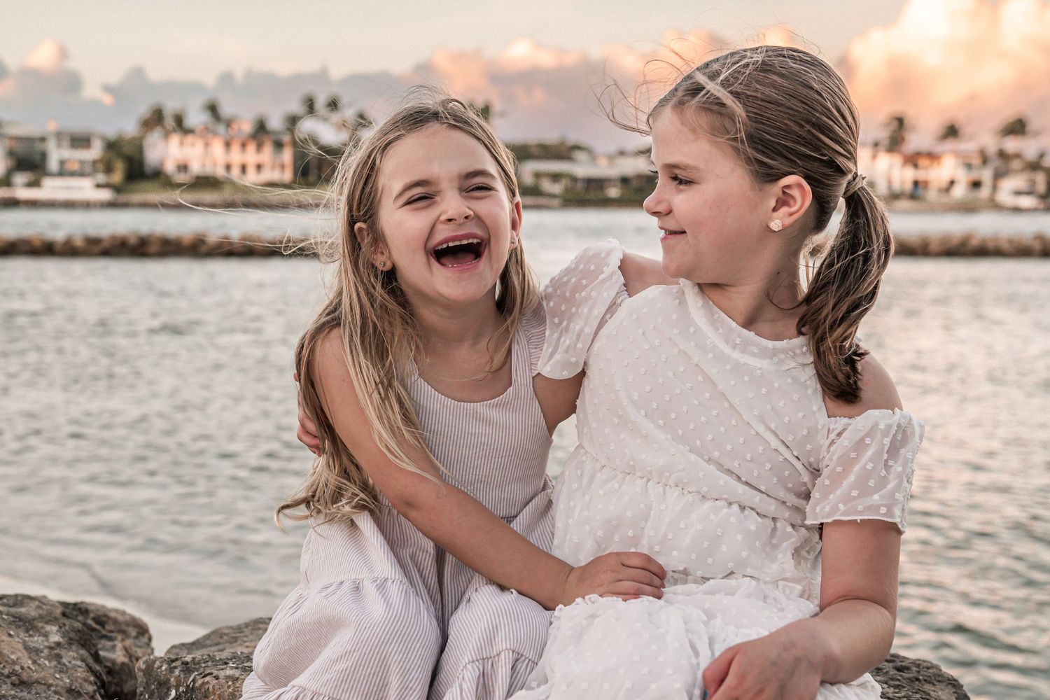Two children in white dresses share a joyful moment on coastal rocks during sunset with buildings in background.