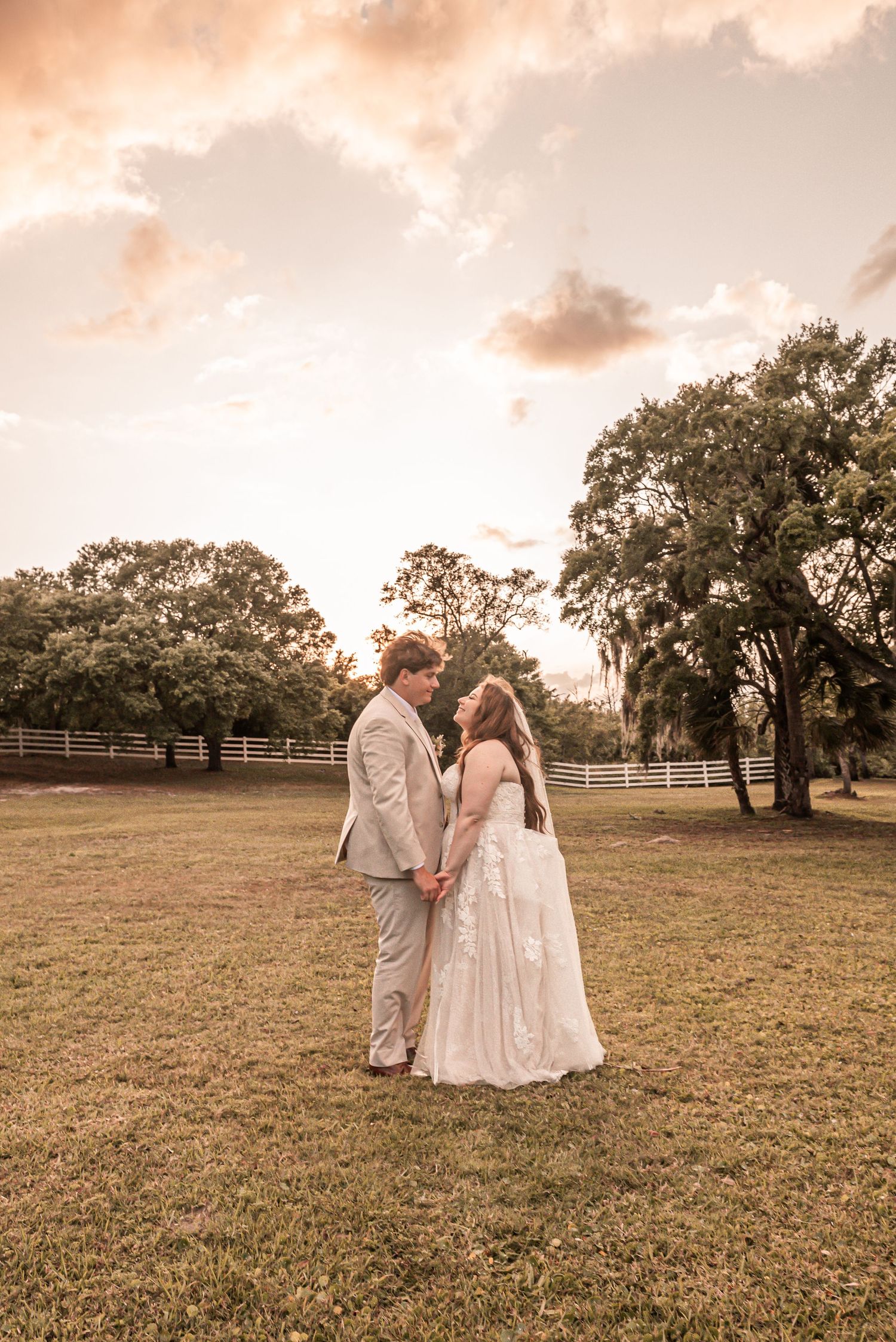 A couple in wedding attire standing in a grassy field at sunset with trees and a fence in the background.