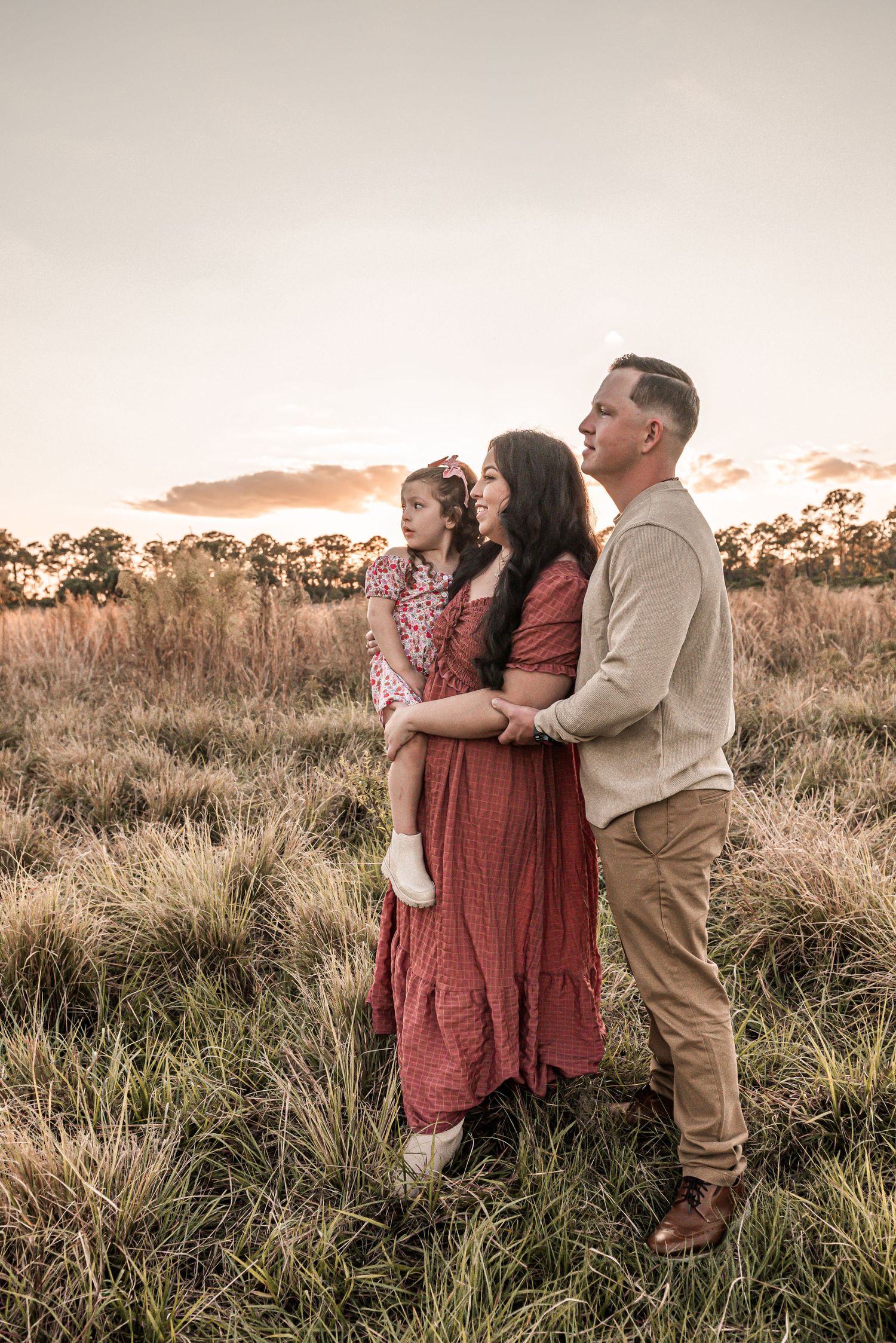 A family embraces in a rustic field at sunset, wearing coordinated outfits in warm tones of burgundy and beige.