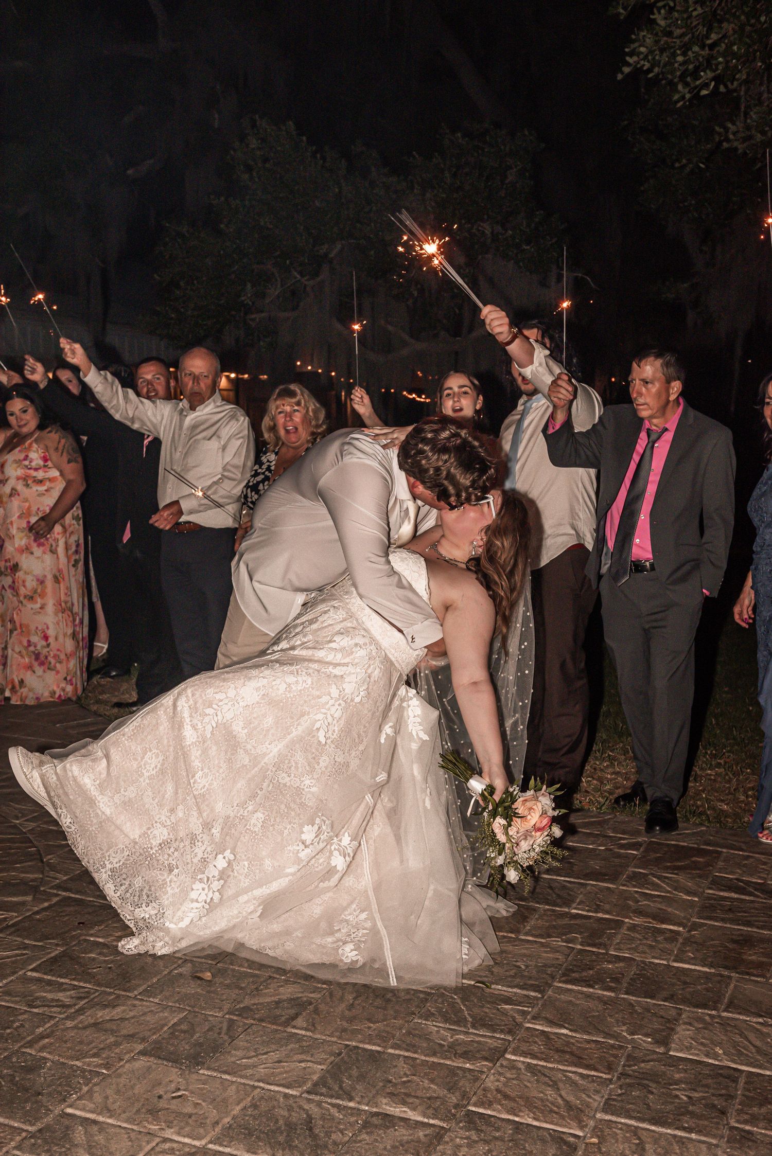 Newlyweds share a romantic dip kiss on a stone patio while wedding guests celebrate with sparklers at night.