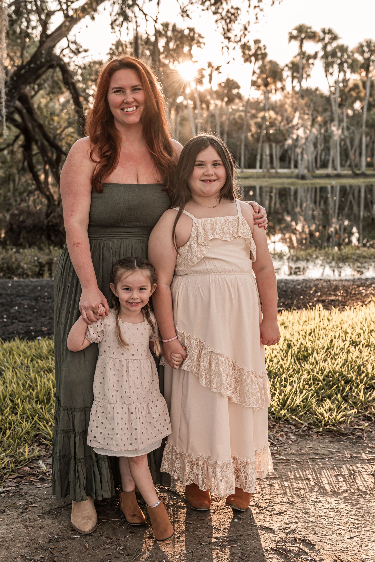 A family portrait at sunset by a lake with three people wearing coordinated dresses in olive and cream tones.