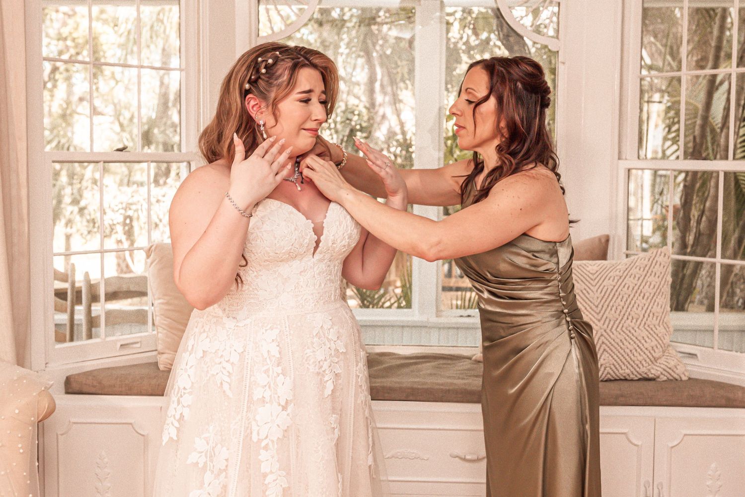 Two women share an emotional moment in a sunlit room as one wearing a metallic dress helps the other in white lace.