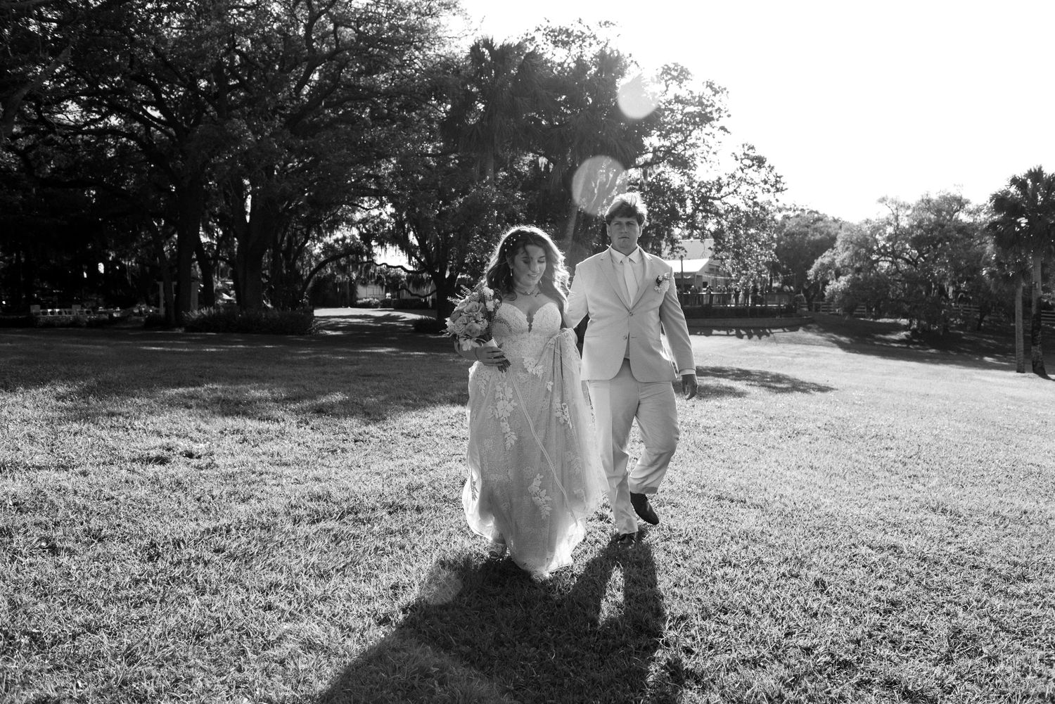 A couple in white wedding attire run joyfully across a sunlit lawn during an outdoor celebration.