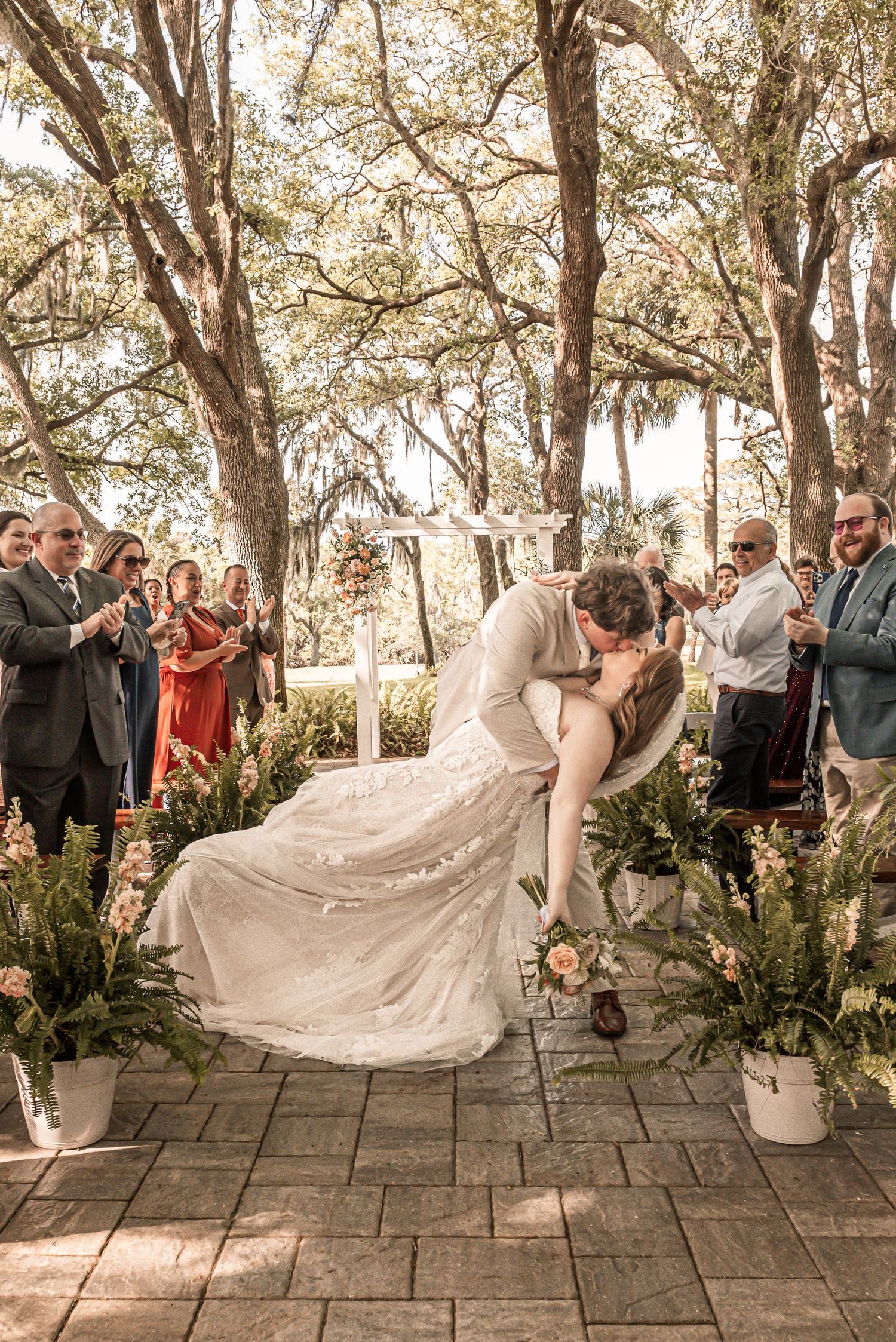 Romantic couple embraces during outdoor wedding ceremony under moss-draped trees with guests watching on stone patio.