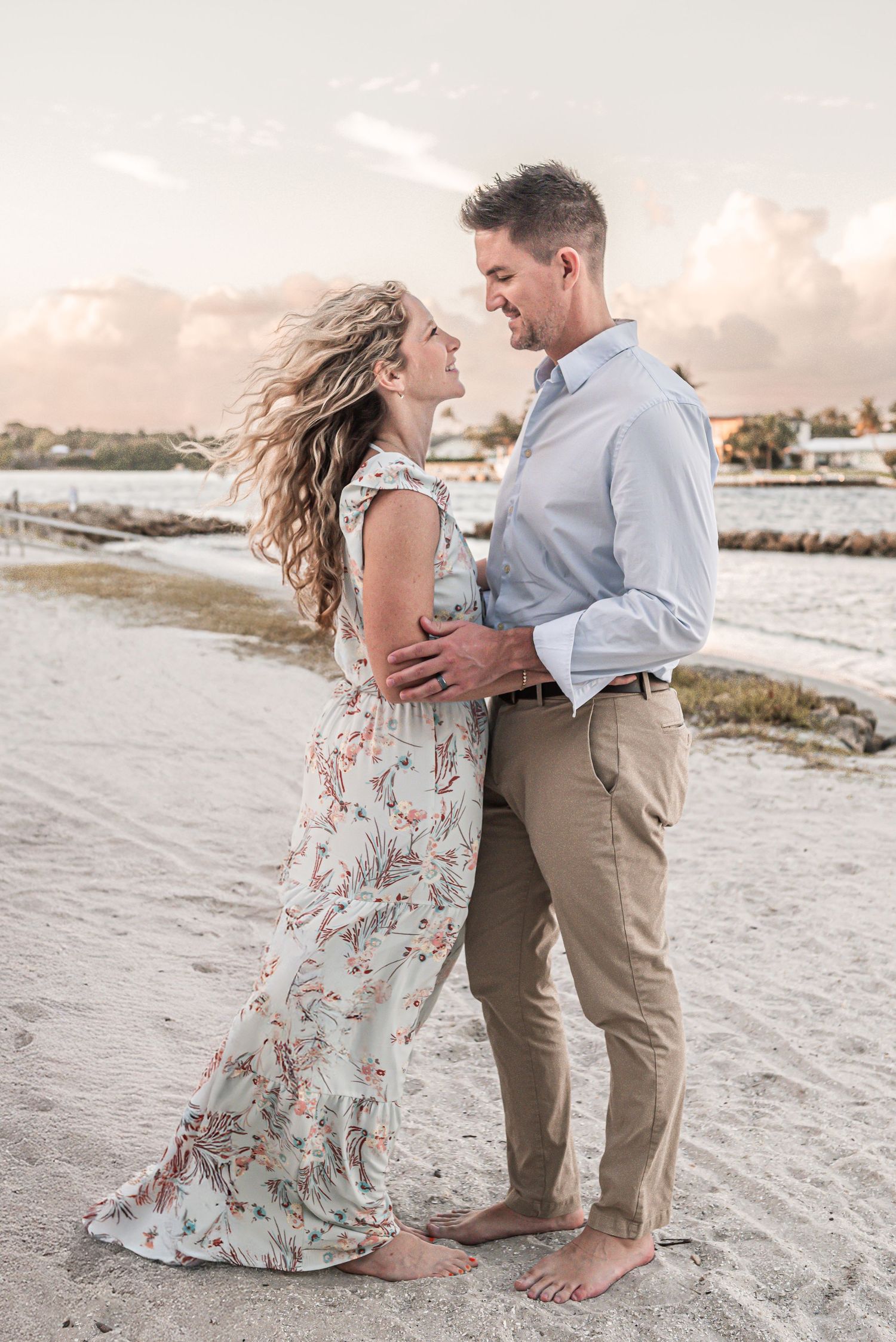 A romantic couple embraces on a sandy beach at sunset wearing flowy dress and casual attire.