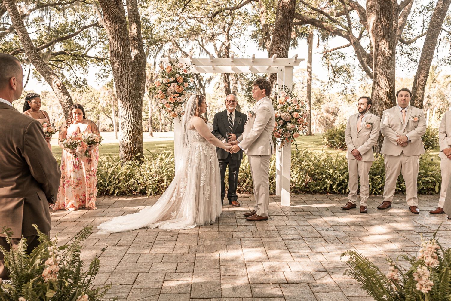 Outdoor garden wedding ceremony taking place on a brick patio surrounded by trees at golden hour.