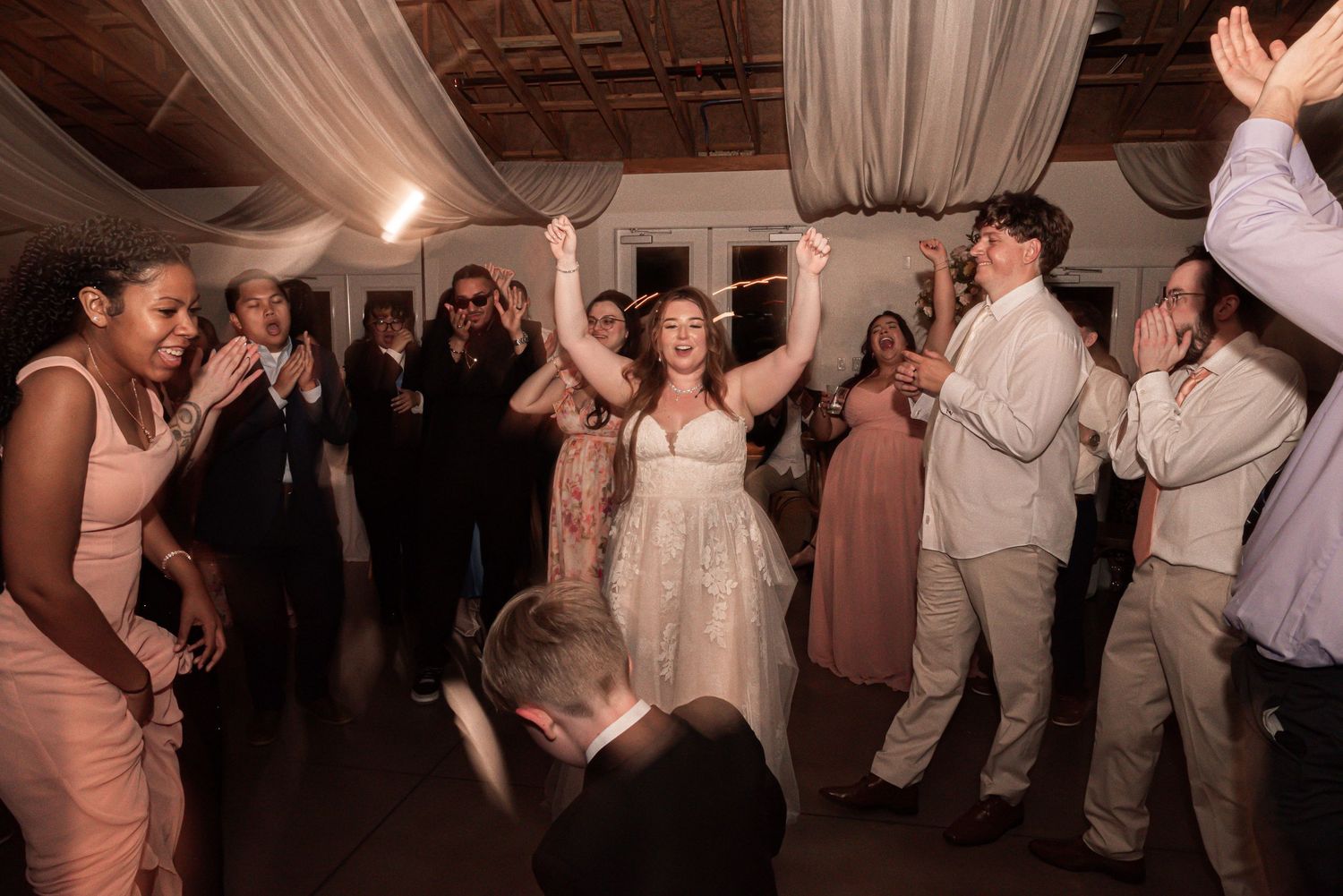 Wedding reception guests dancing and celebrating on the dance floor at an indoor venue decorated with draped fabric.