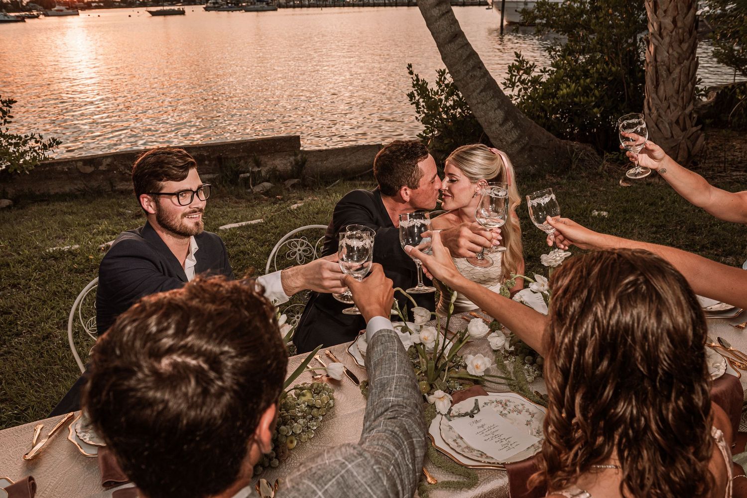 Group of friends toasting with wine glasses at outdoor lakeside gathering during sunset.